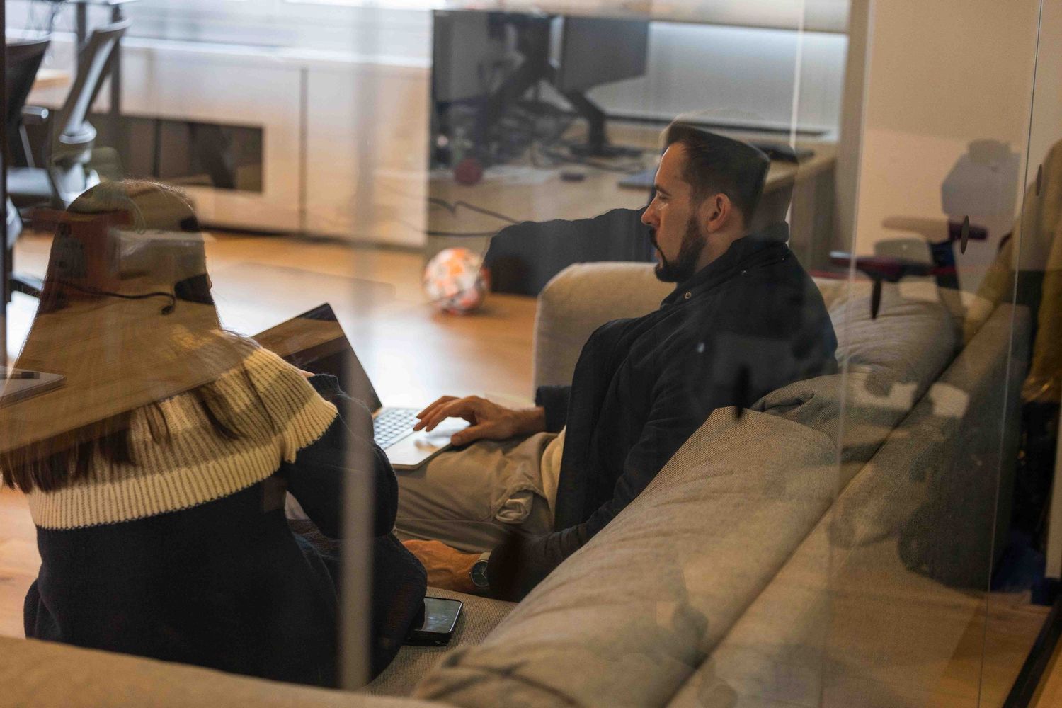 looking through a glass window where a man and female colleague are talking on a sofa