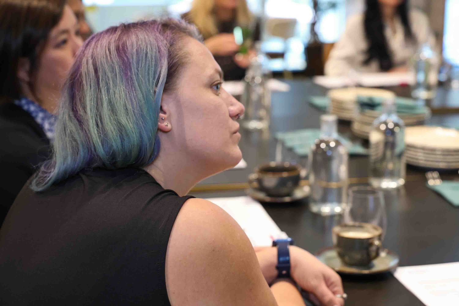 A close up of a women with purple hair looking at someone speaking at a meeting