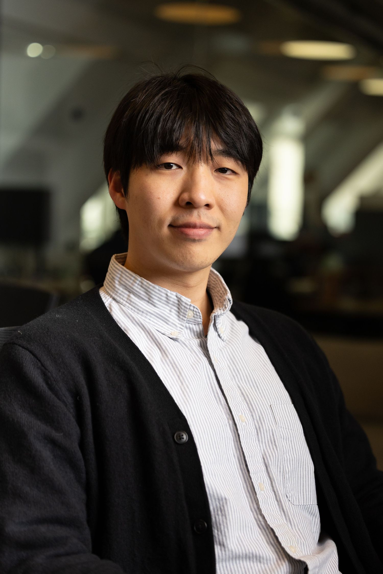A head shot of a Asian man in white shirt and black cardigan with the office behind him
