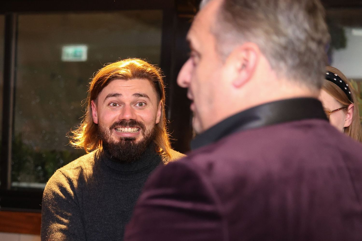 A man with a beard and long hair smiling over the shoulder of another man at an event