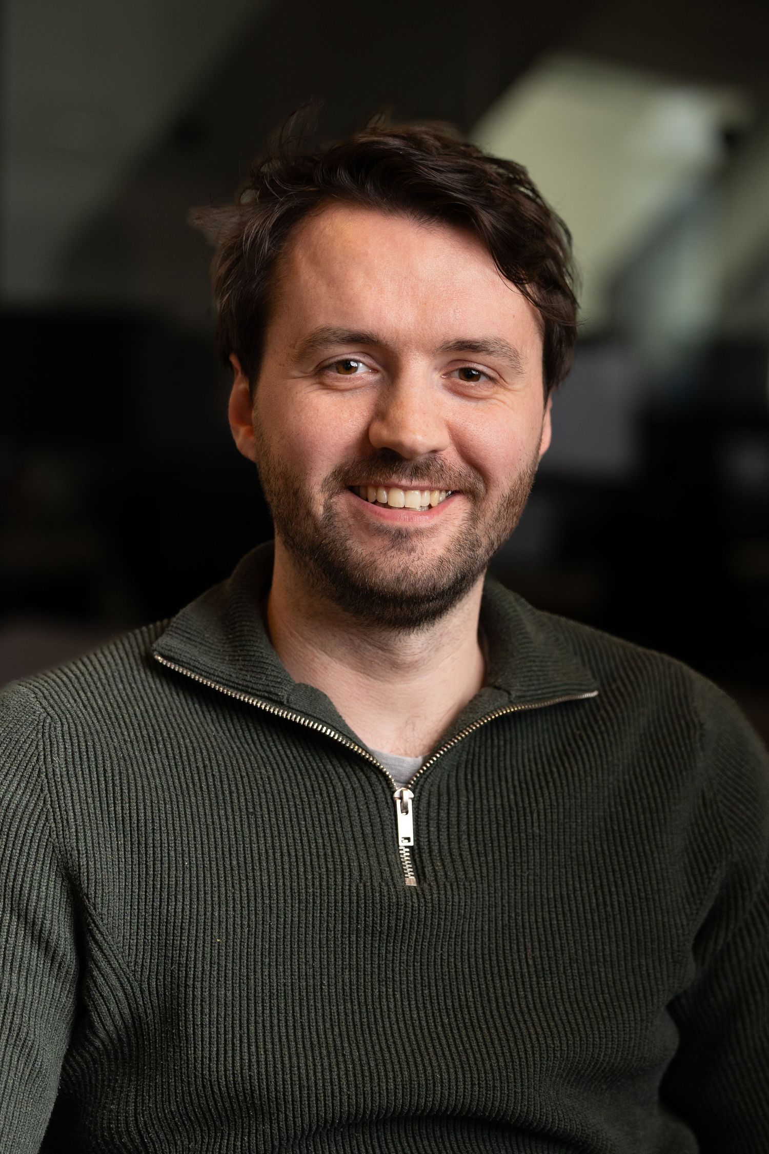 A head shot of a white man with a green jumper on with the office behind him