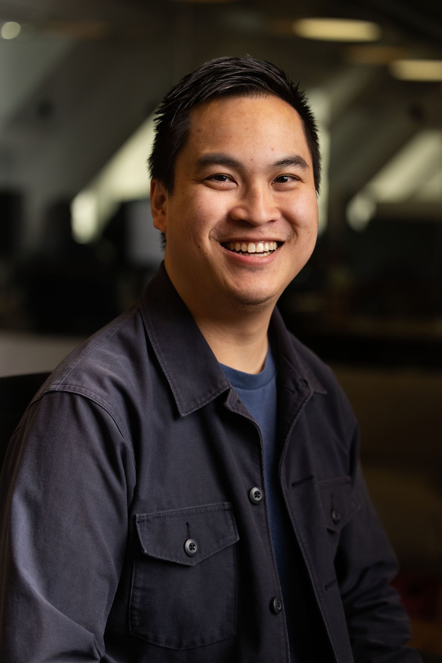 A head shot of a Asian man with a blue tshirt with the office behind him