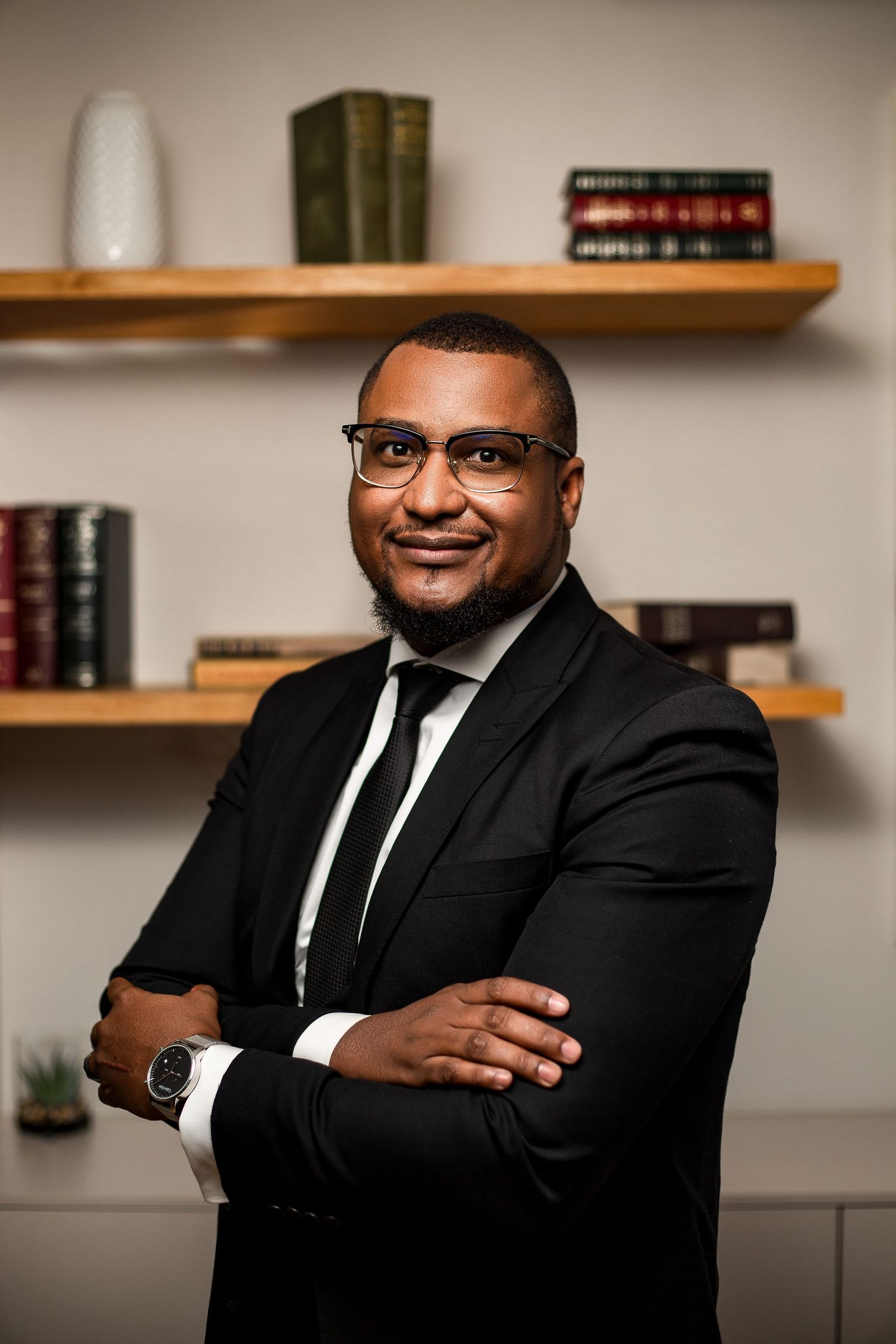 Professional headshots of a person in a black suit and tie against a neutral office background with bookshelves.