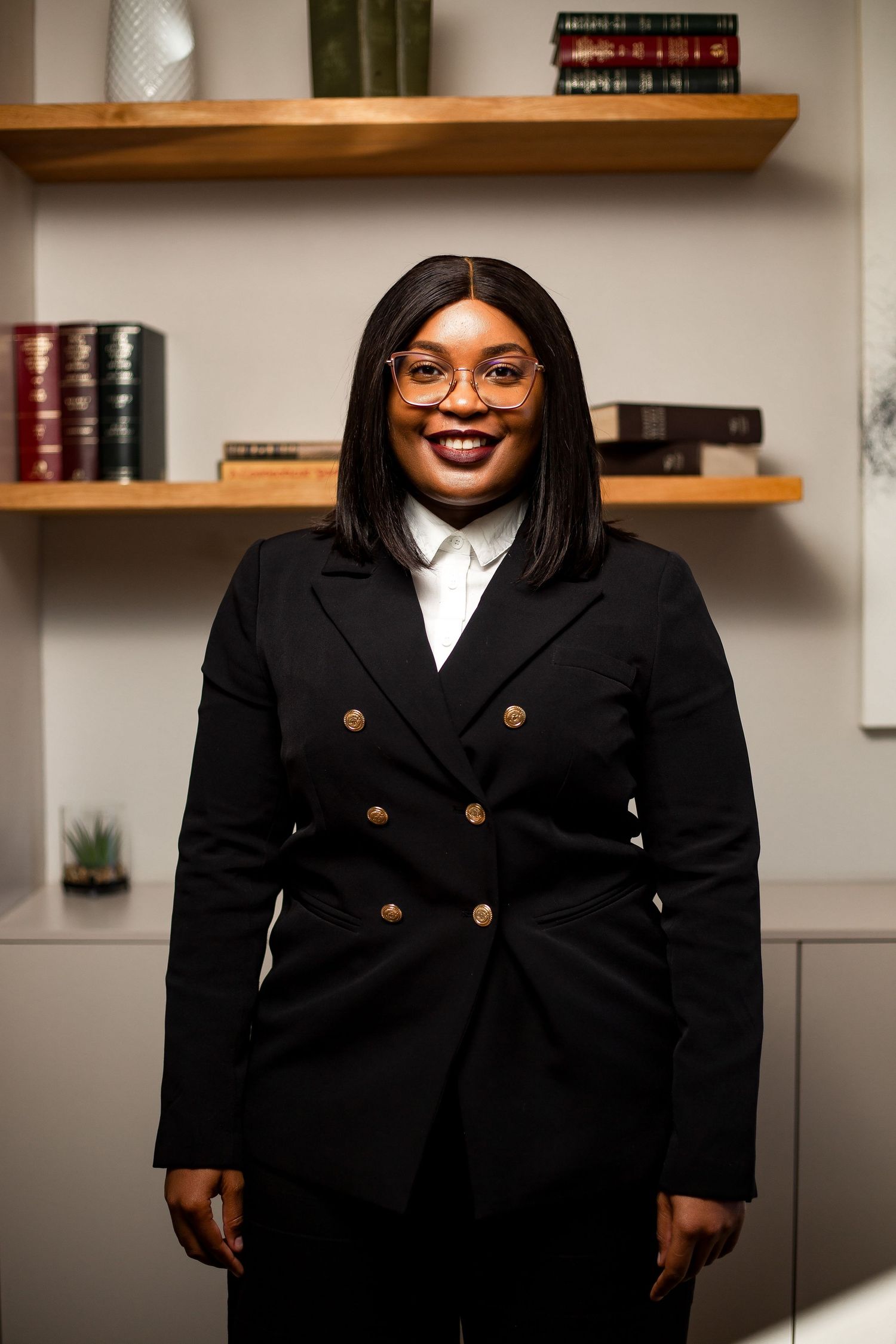 Professional headshot of person in black blazer with glasses against office background.