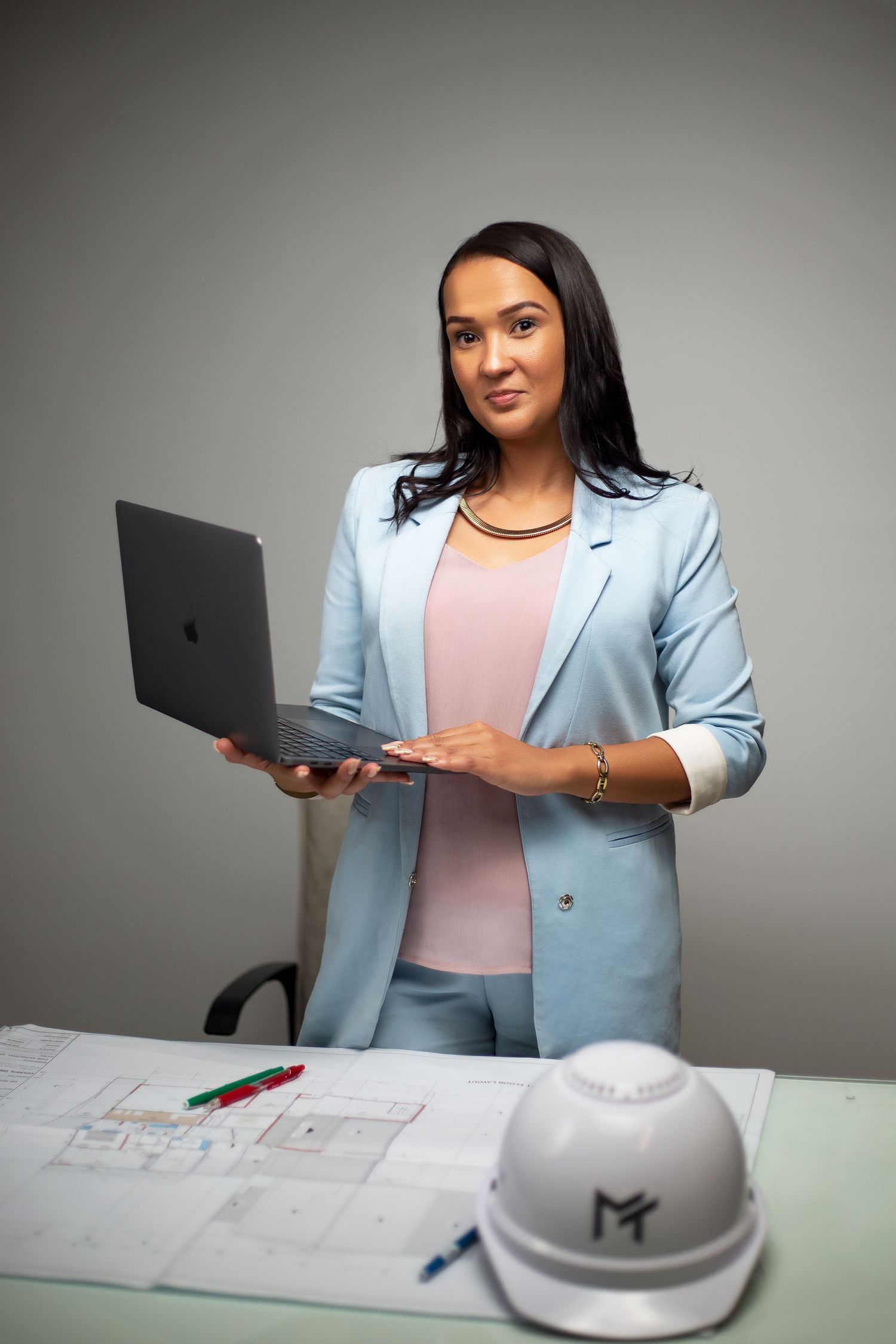 Architect or engineer in light blue blazer reviews plans while holding laptop with hard hat on desk.