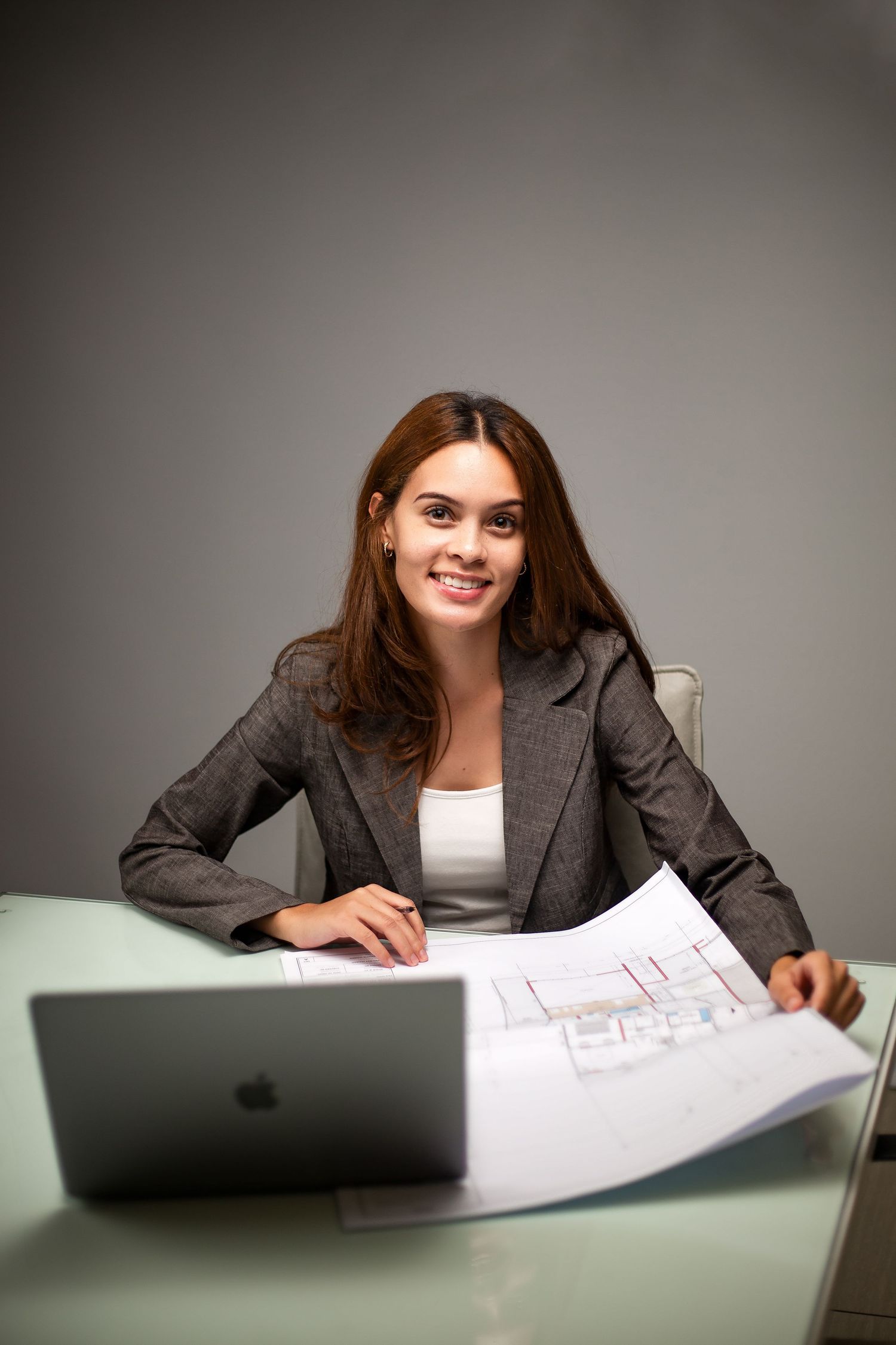 Professional woman in business attire working on blueprints with laptop at desk in modern office setting.