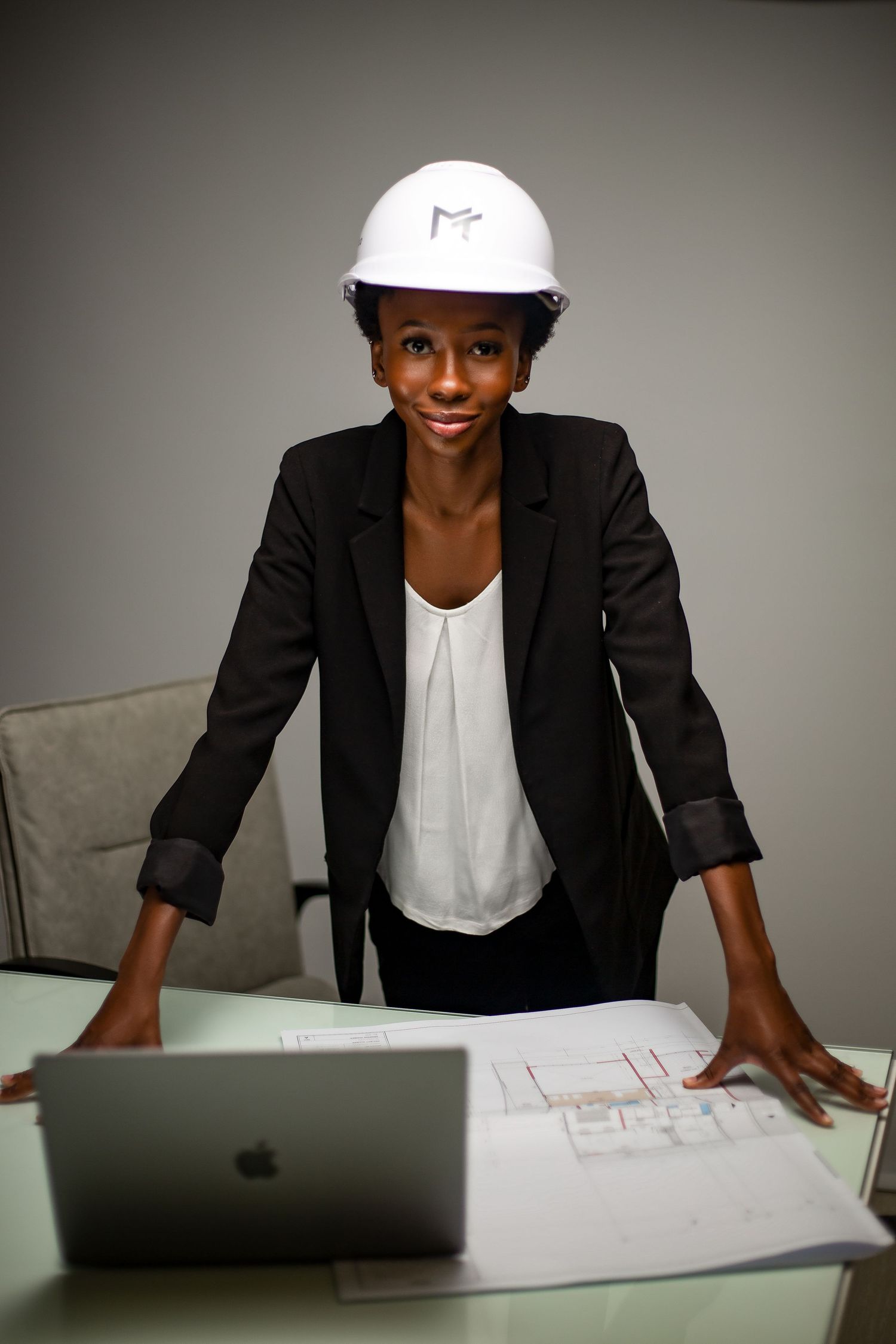 Professional at work wearing hardhat and blazer reviewing documents at desk with laptop.