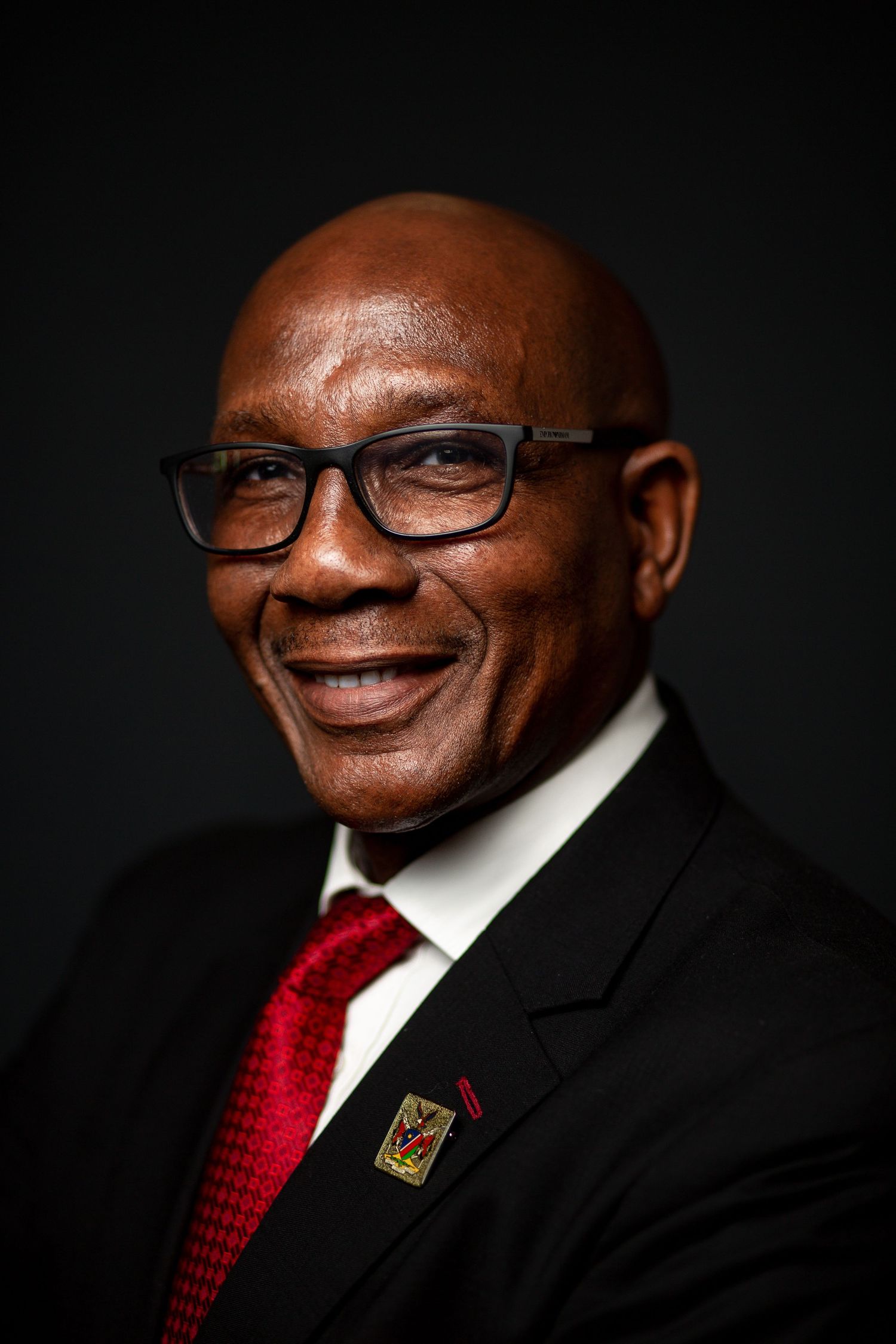 Close-up professional headshots showing warm expressions with glasses and red tie against dark backdrop.