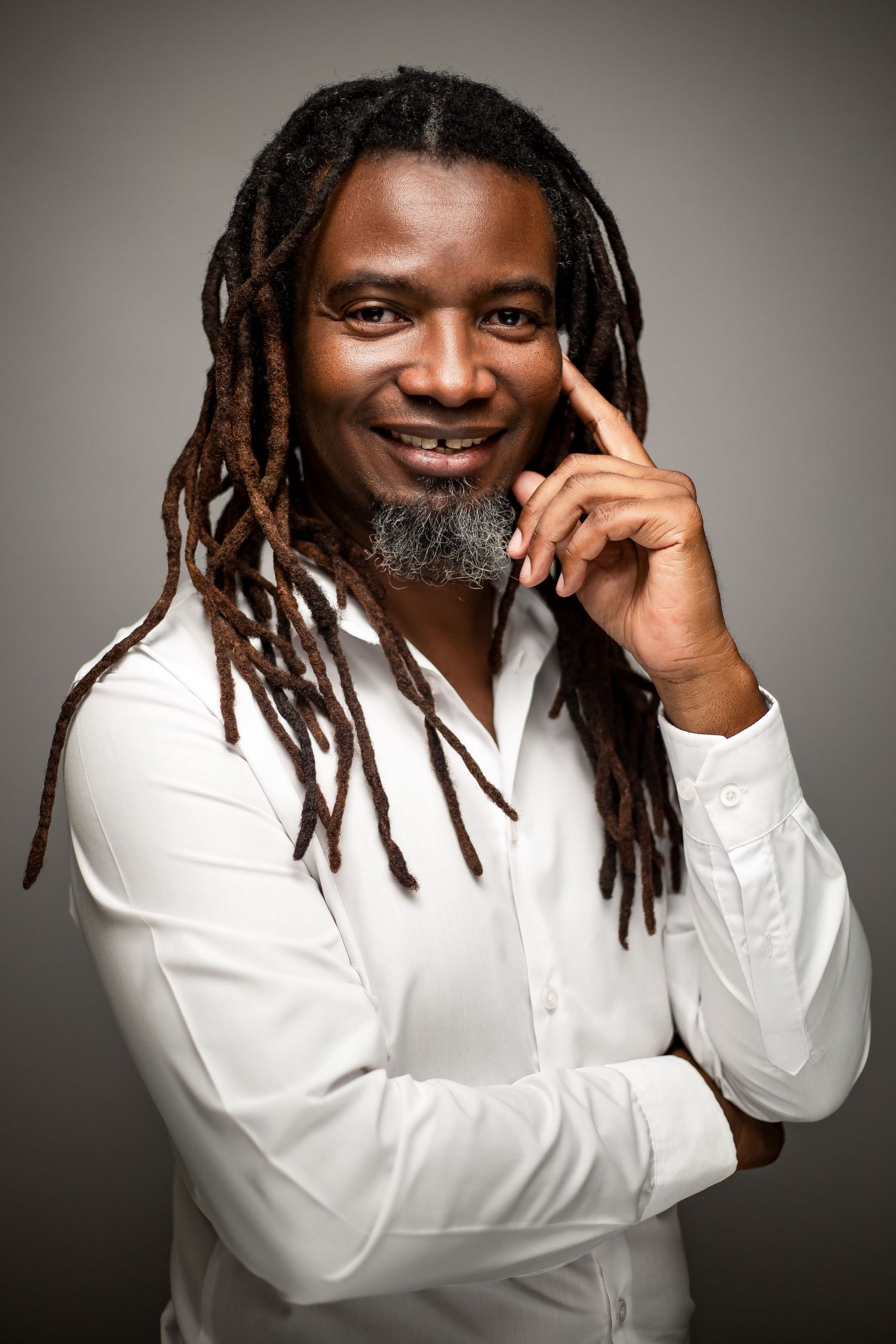 Professional in white shirt with dreadlocks poses thoughtfully against gray background.