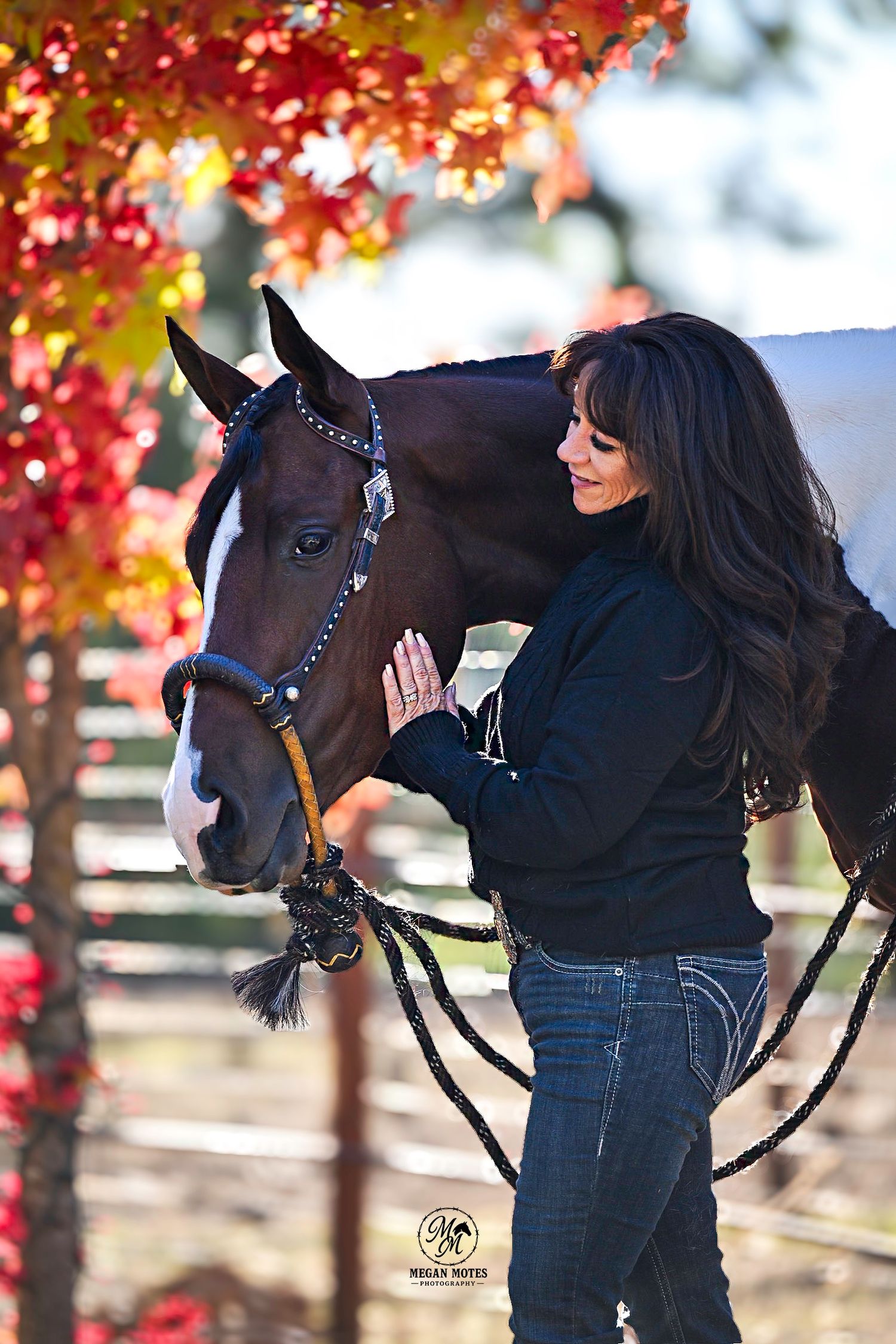 Woman in black jacket gently touches a chestnut horse wearing a blue halter, surrounded by vibrant red autumn foliage.