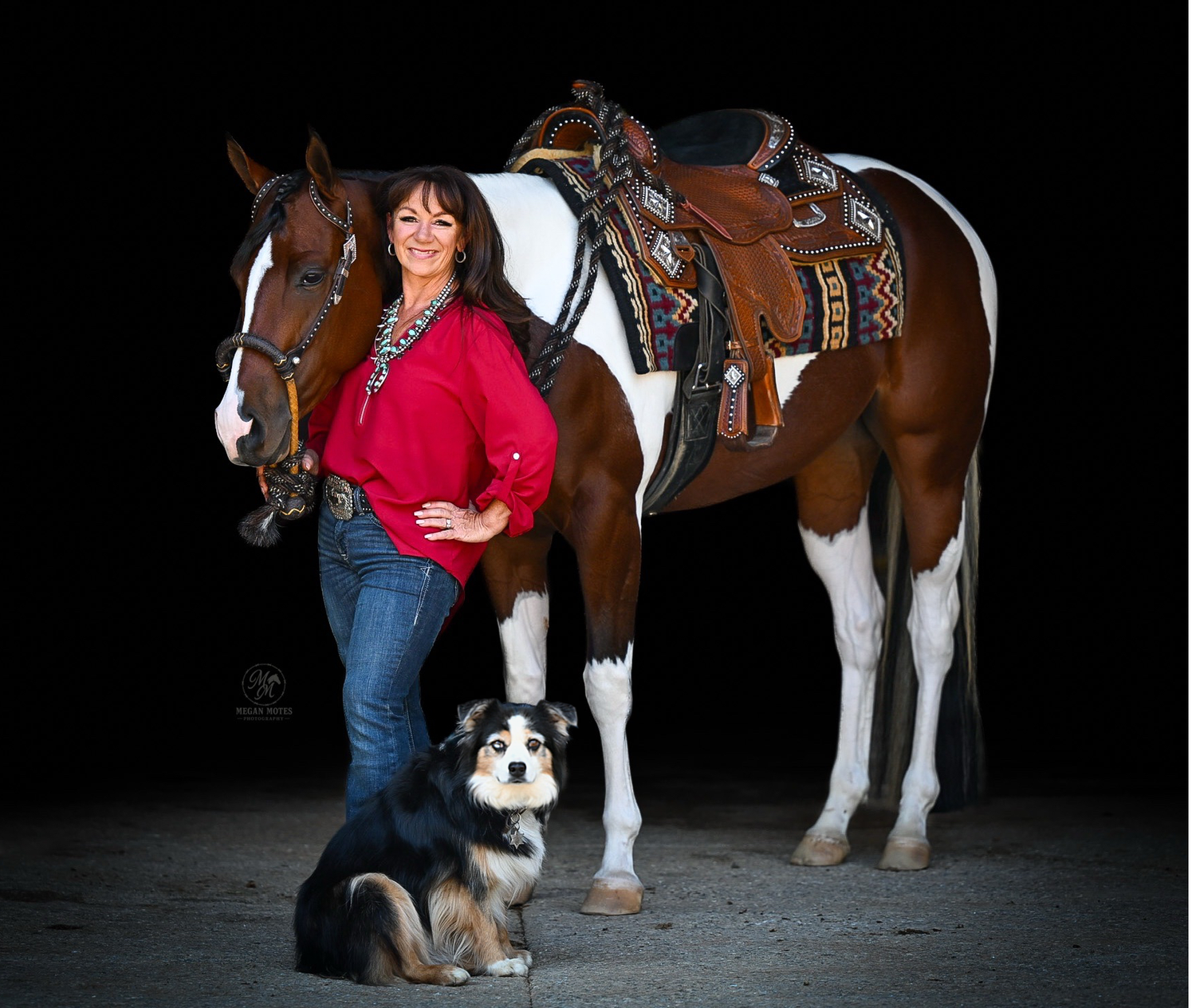 Woman in red shirt poses with a saddled Paint horse and Australian Shepherd dog against black background.