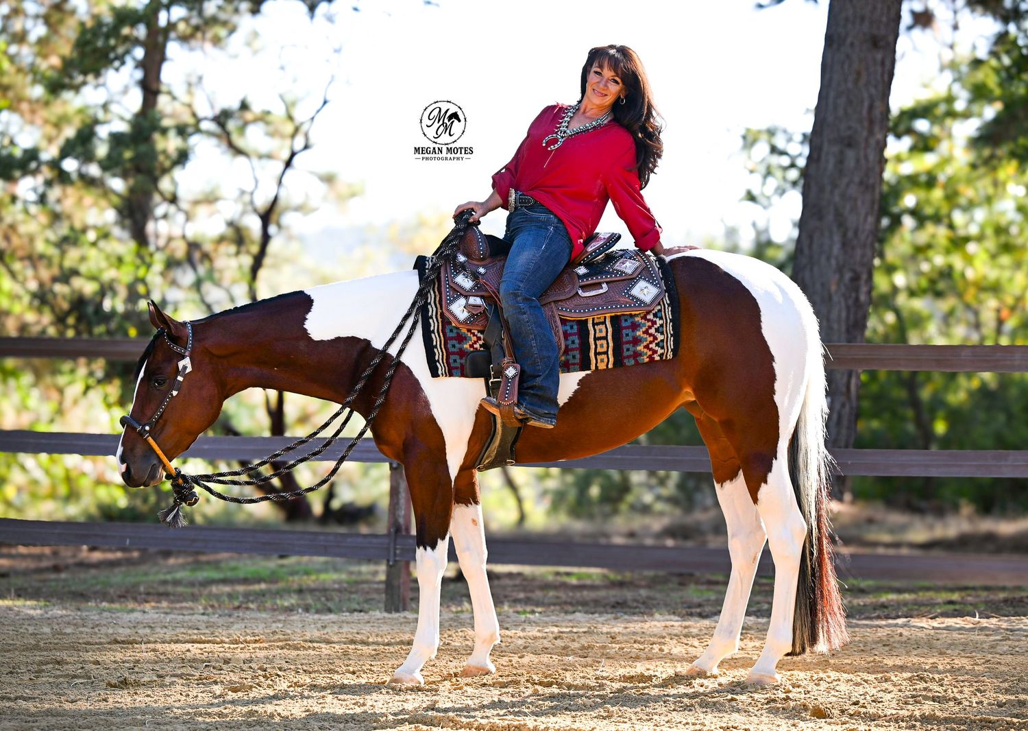 Woman in red shirt riding a brown and white paint horse outdoors in a rustic equestrian setting.