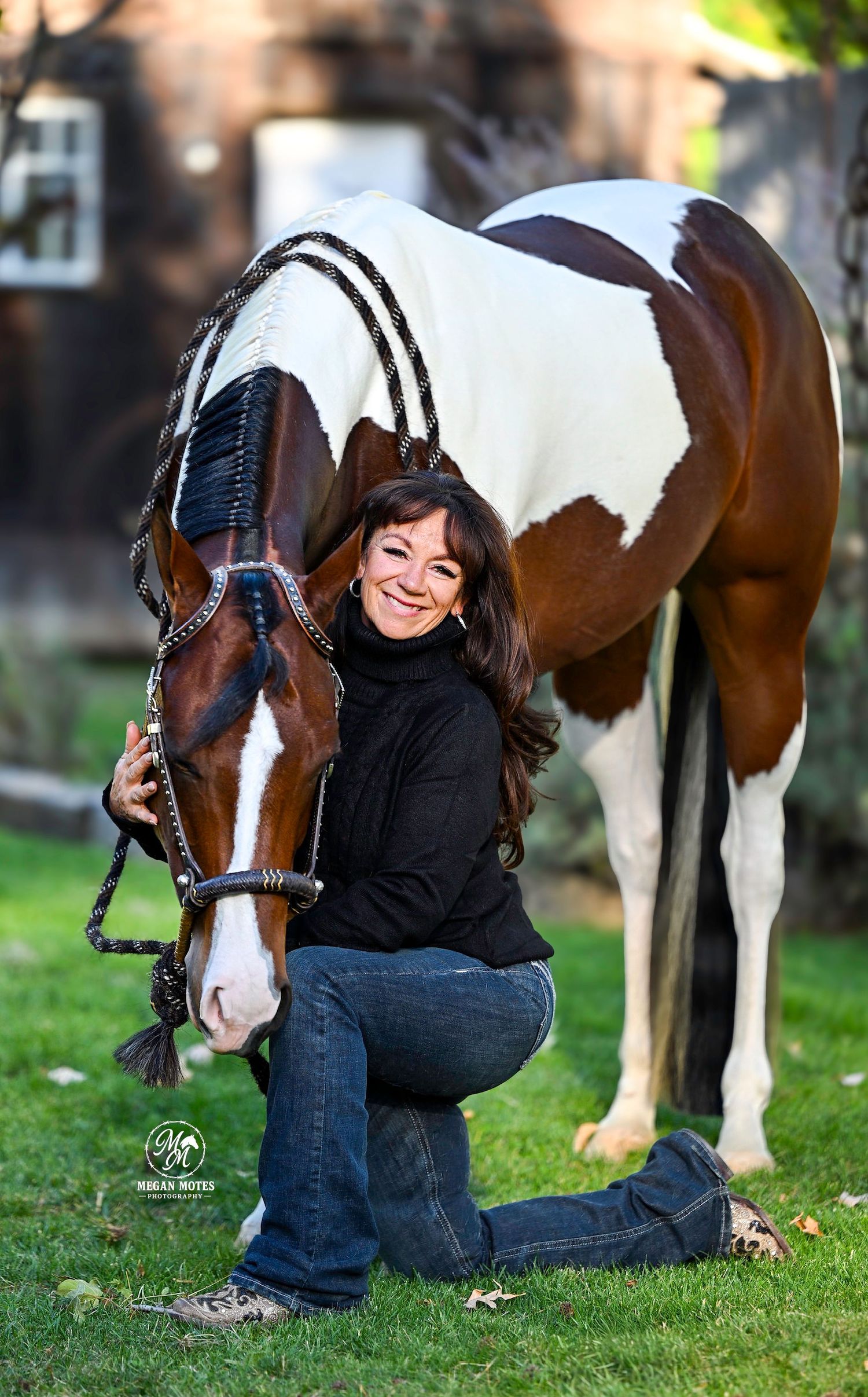 Woman kneeling beside a brown and white paint horse with braided mane on green grass outdoors.