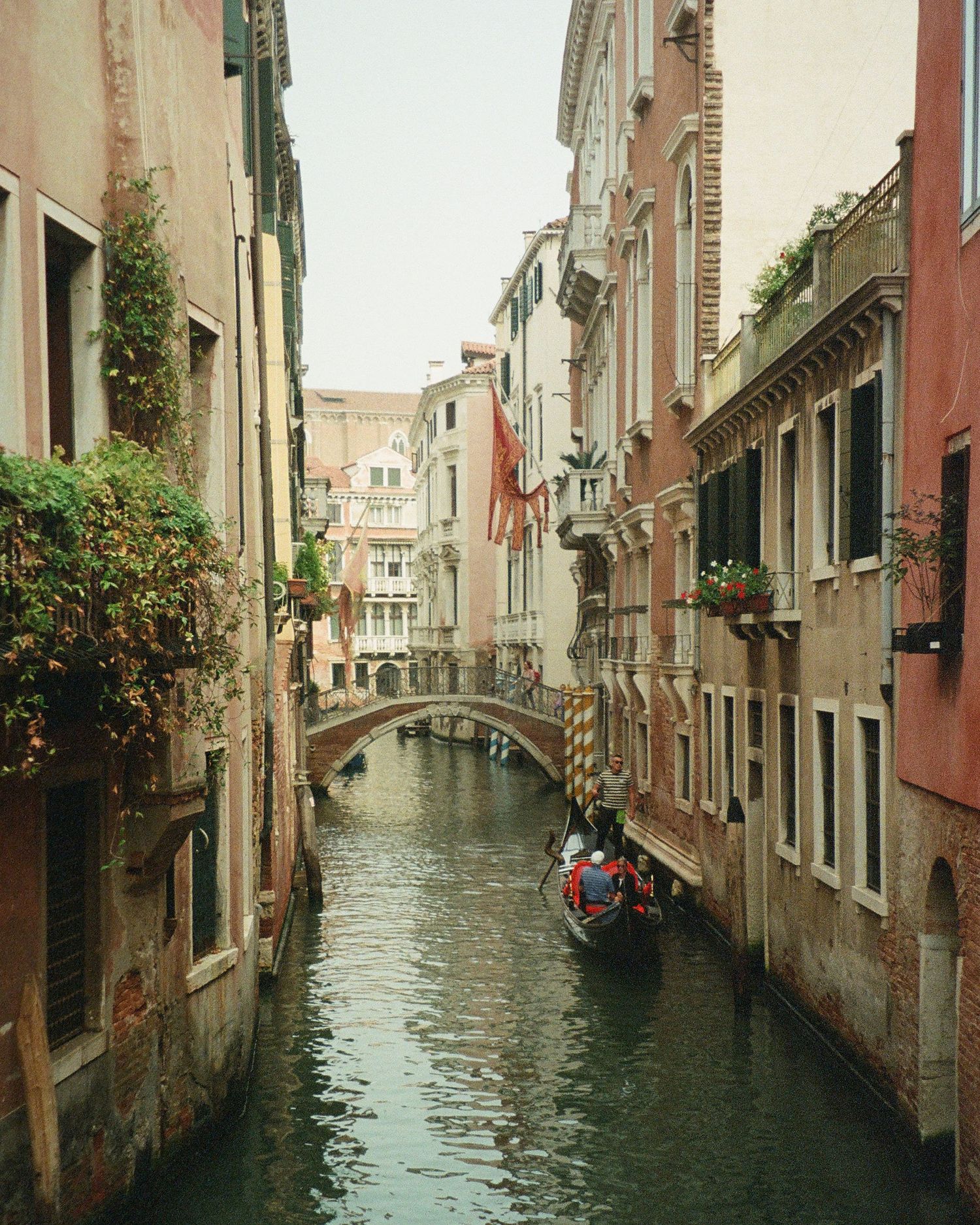 Film photograph of narrow canal in Venice with historic buildings and gondolas reflecting in the calm water during daytime.
