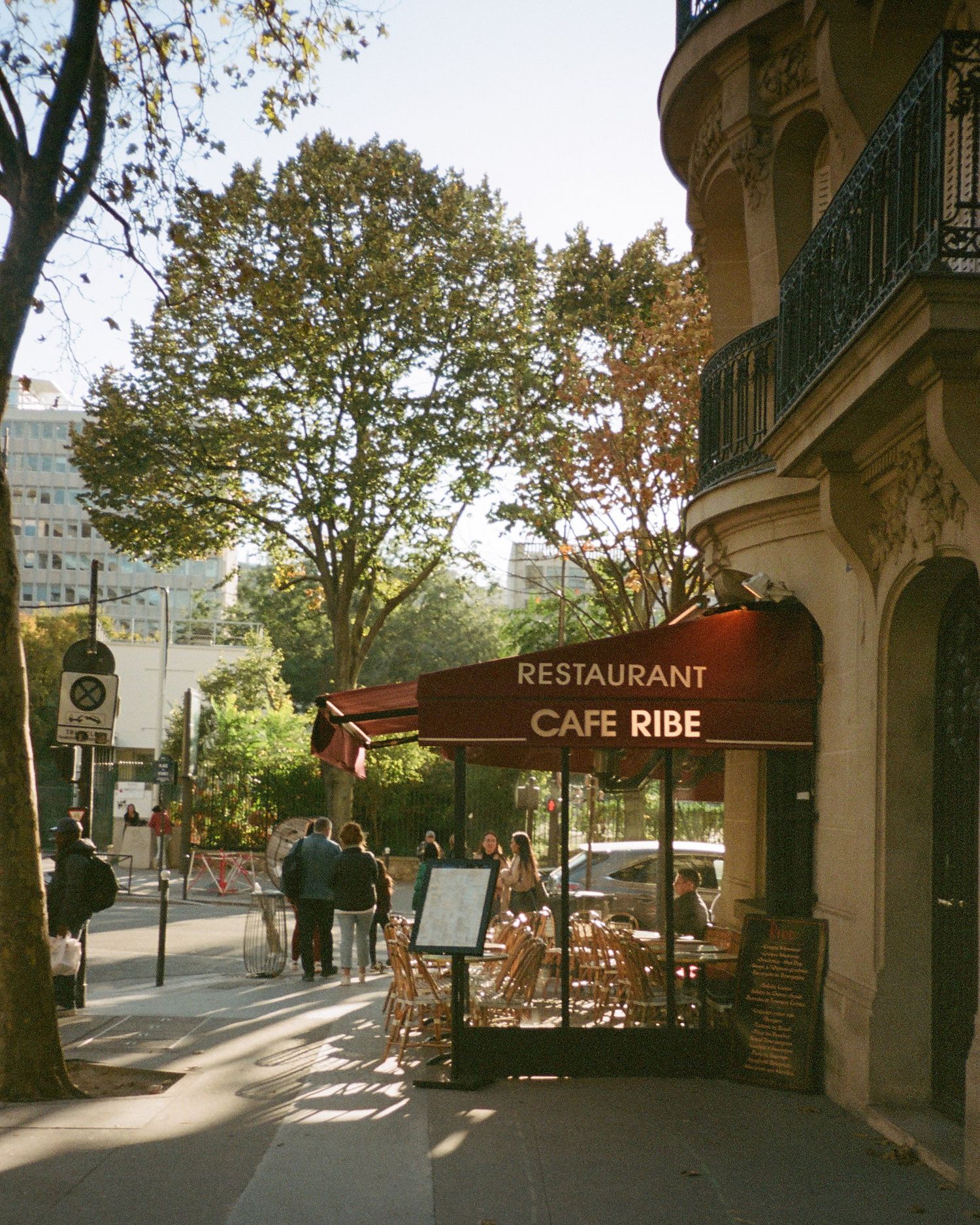 Film photograph of a charming Parisian cafe with outdoor seating and wrought iron balconies on a tree-lined street during golden hour.