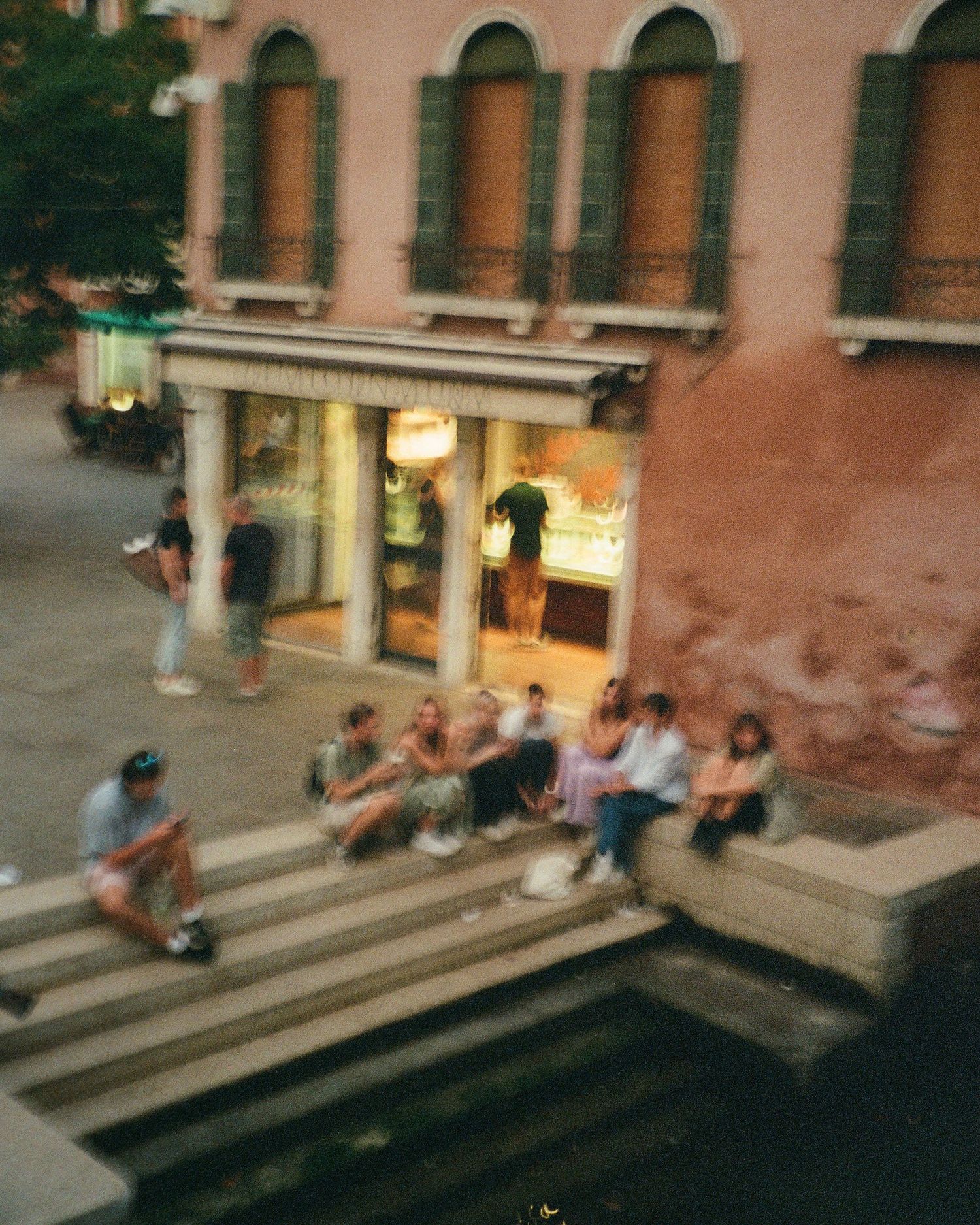 Film photograph of people sitting on steps outside an illuminated storefront with arched windows in Venice at dusk.