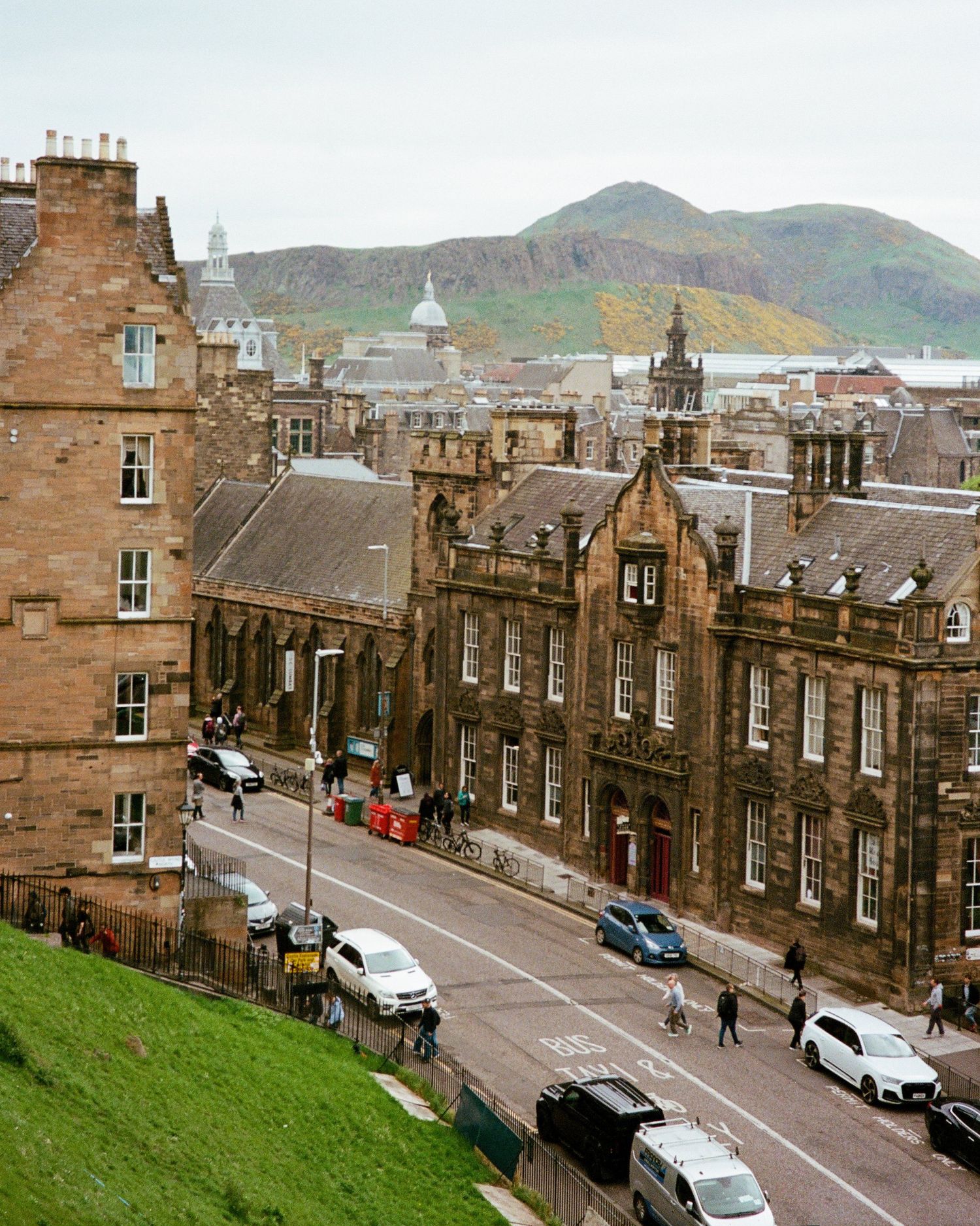 Film photograph of historic stone buildings lining a sloping street in Edinburgh's Old Town with Arthur's Seat visible in the background.