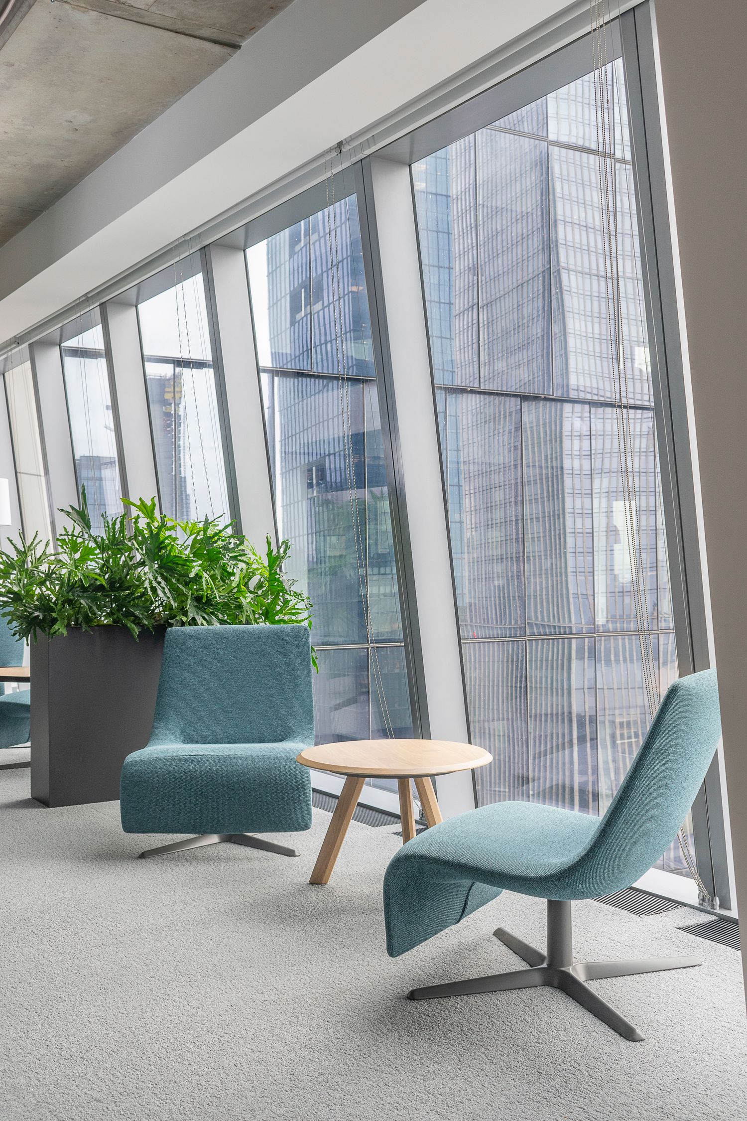 Stylish office lounge with curved teal chairs, wood table, and large windows showcasing cityscape and natural daylight, alongside greenery.