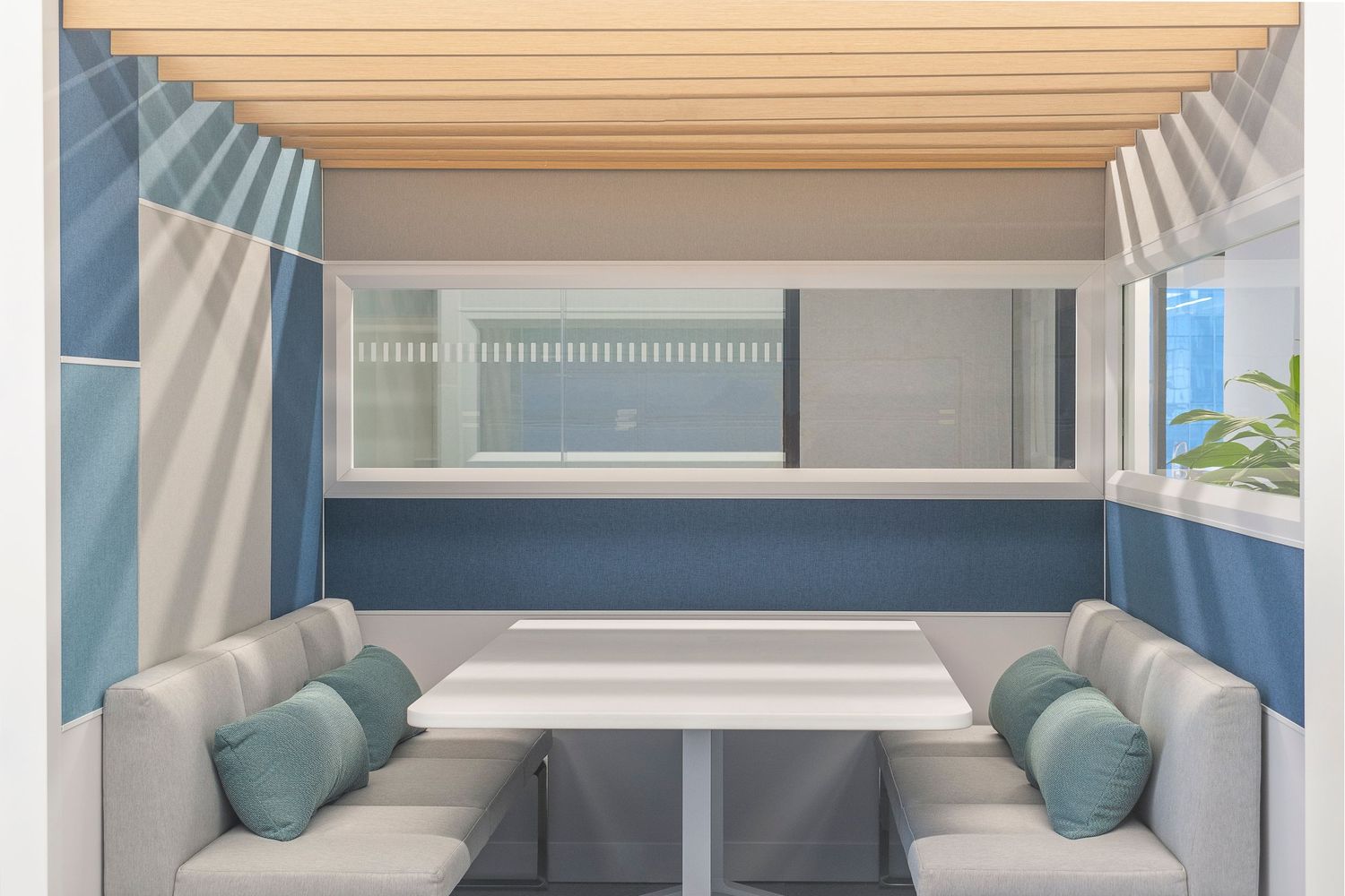 Private work booth with soft gray seating, accent pillows, blue and beige wall panels, and wood slatted ceiling in a modern office setting.