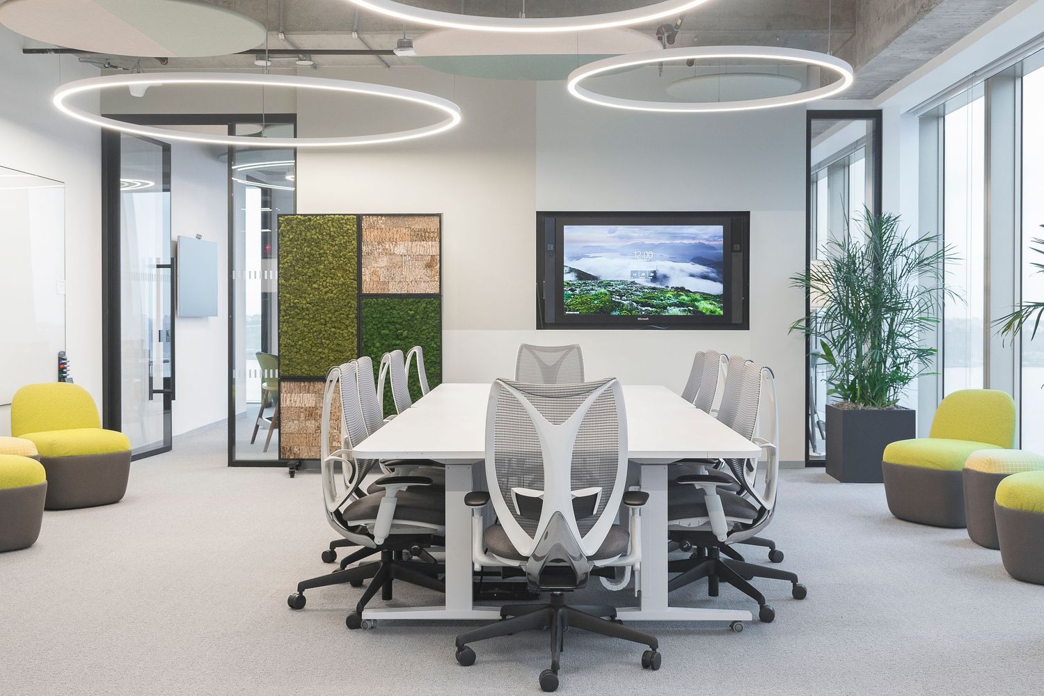 Contemporary office conference room with ergonomic mesh chairs, biophilic wall panels, digital display, and floor to ceiling windows.