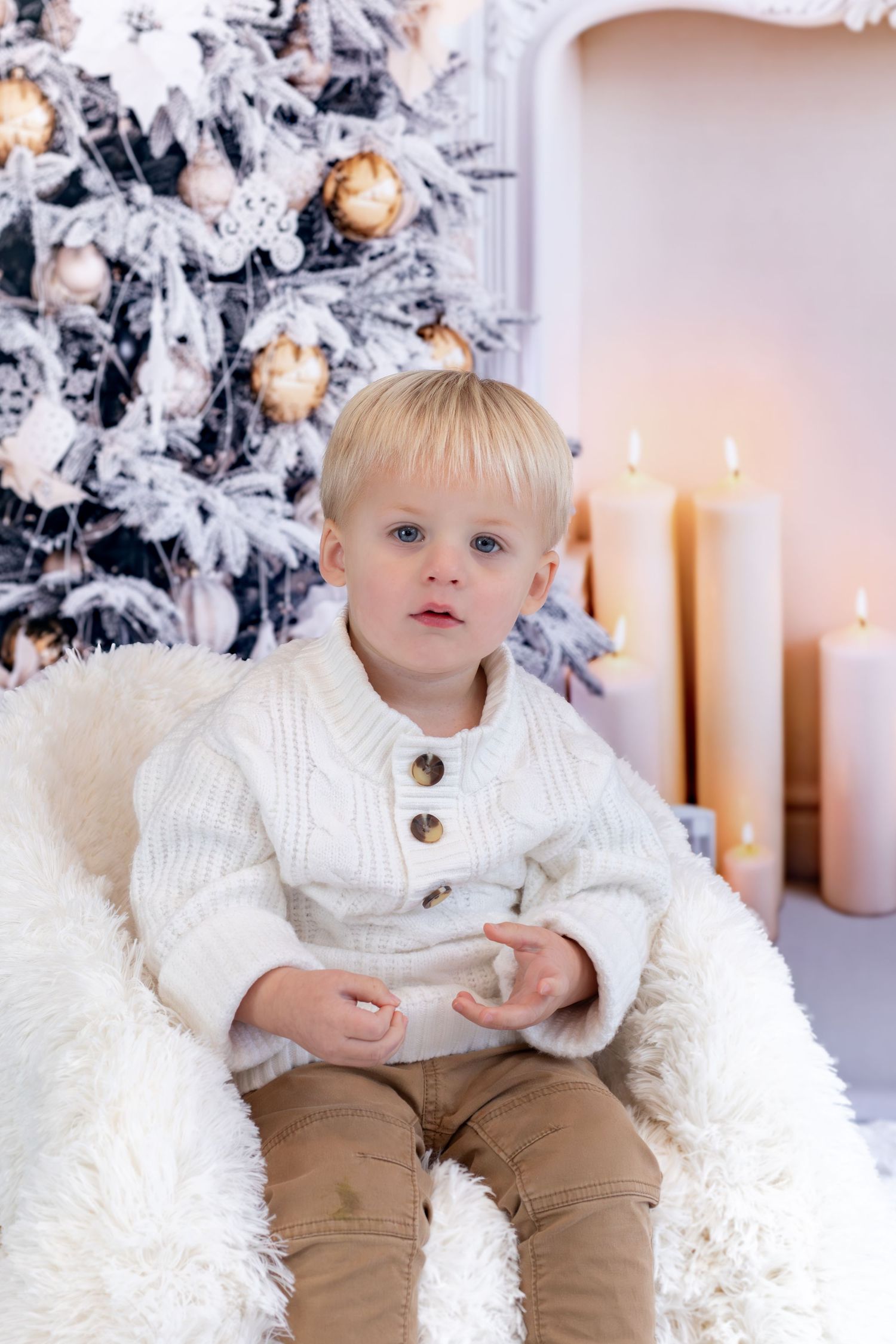 A child in a white knit sweater and brown pants sits by a snowy Christmas tree and lit candles.