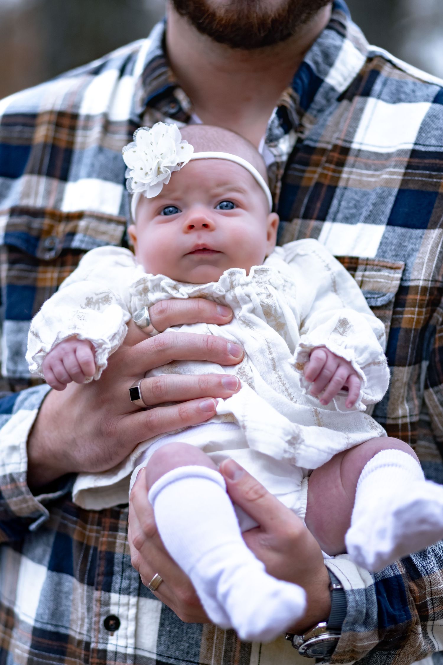 A baby wearing a white lace dress and headband is held by someone in a plaid flannel shirt.
