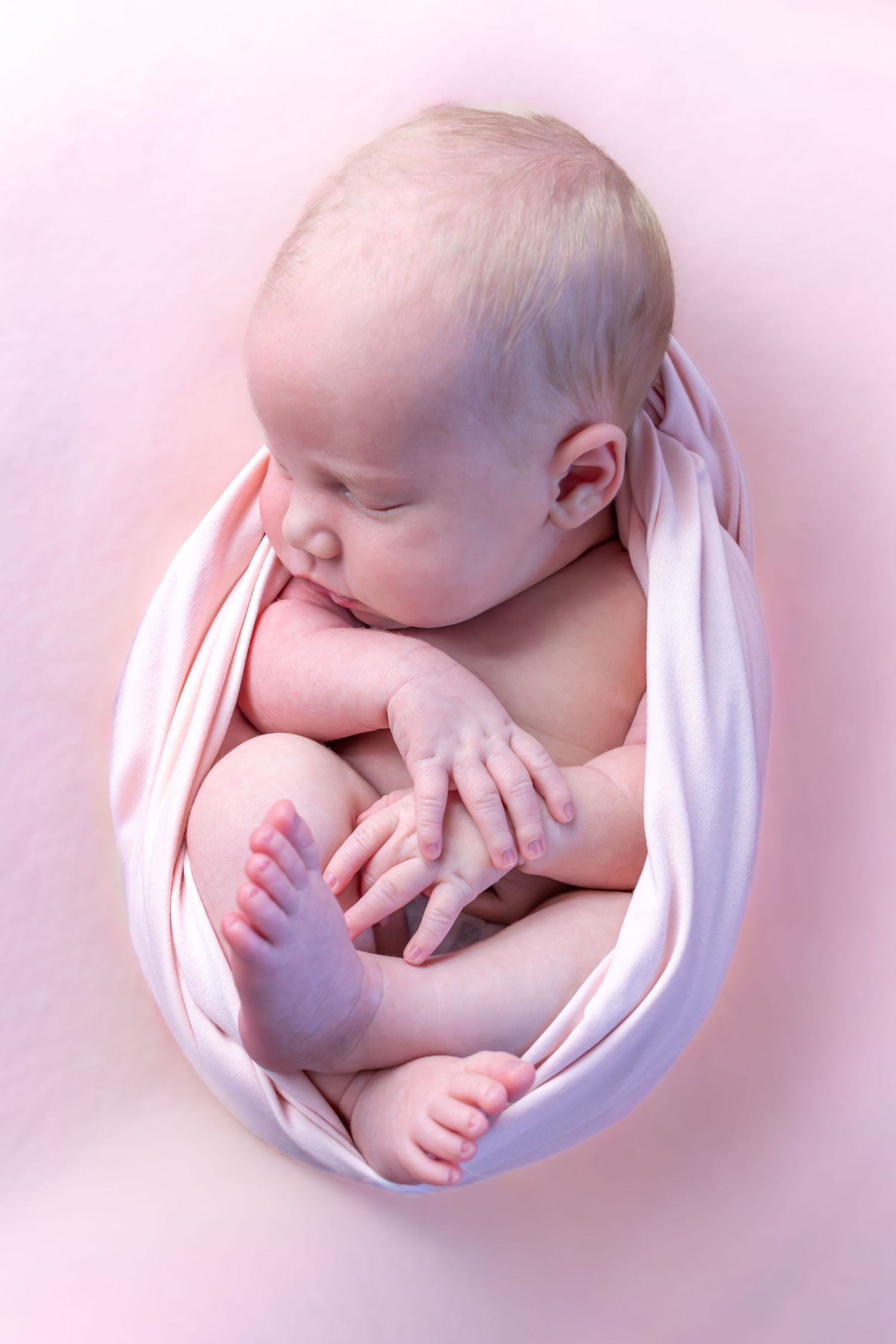 Newborn baby wrapped in white fabric against pink background in artistic pose.