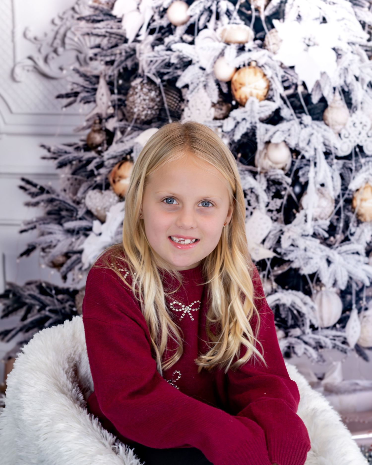 A child in a burgundy sweater poses in front of a snow-covered Christmas tree and white decor.