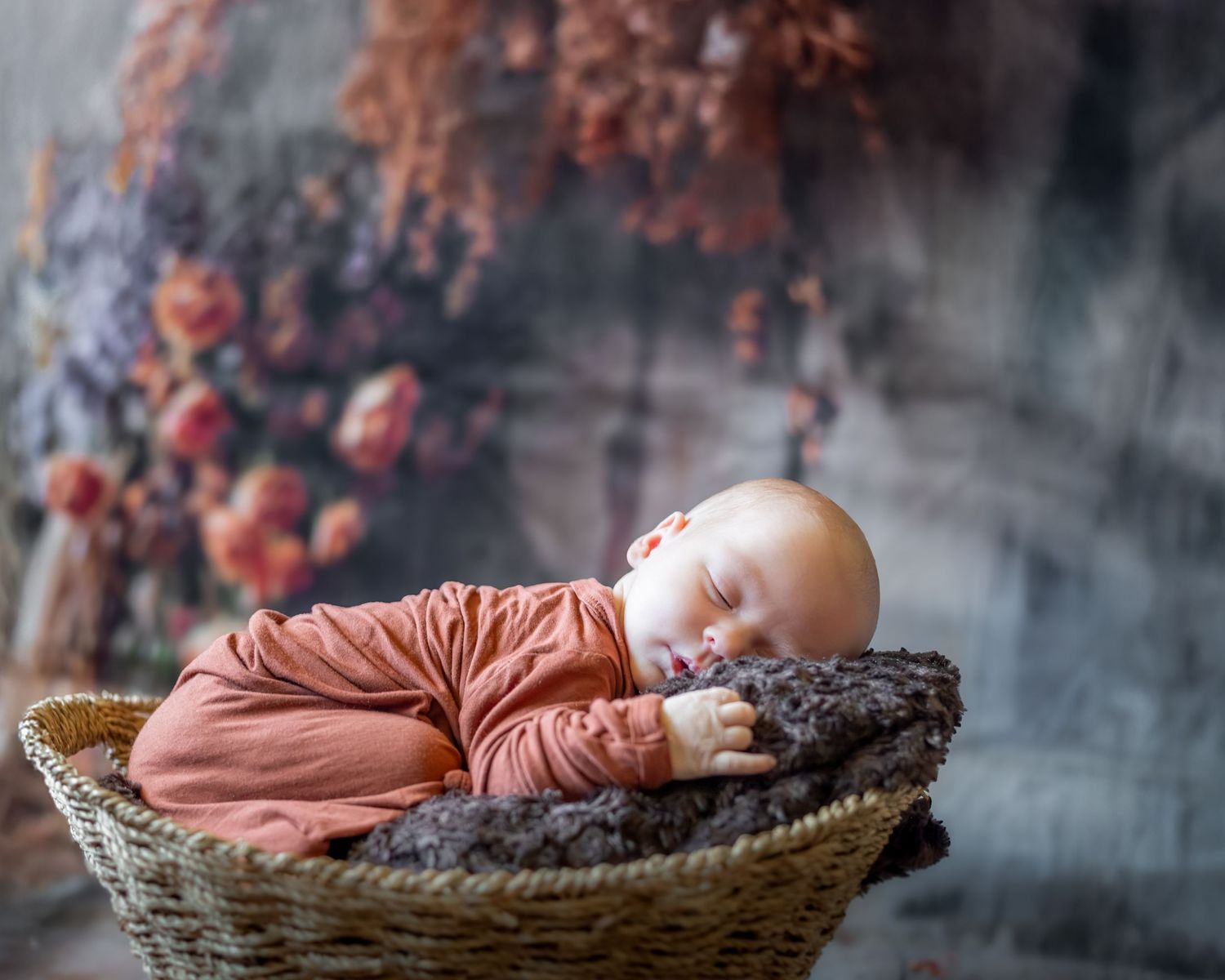 A sleeping baby in a salmon-colored onesie rests peacefully in a woven basket against a dreamy autumn backdrop.