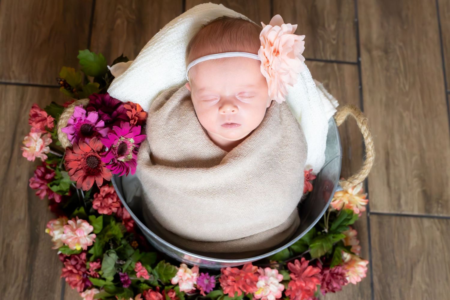 Sleeping newborn wrapped in beige blanket in metal bowl surrounded by pink and purple flowers on wooden floor.