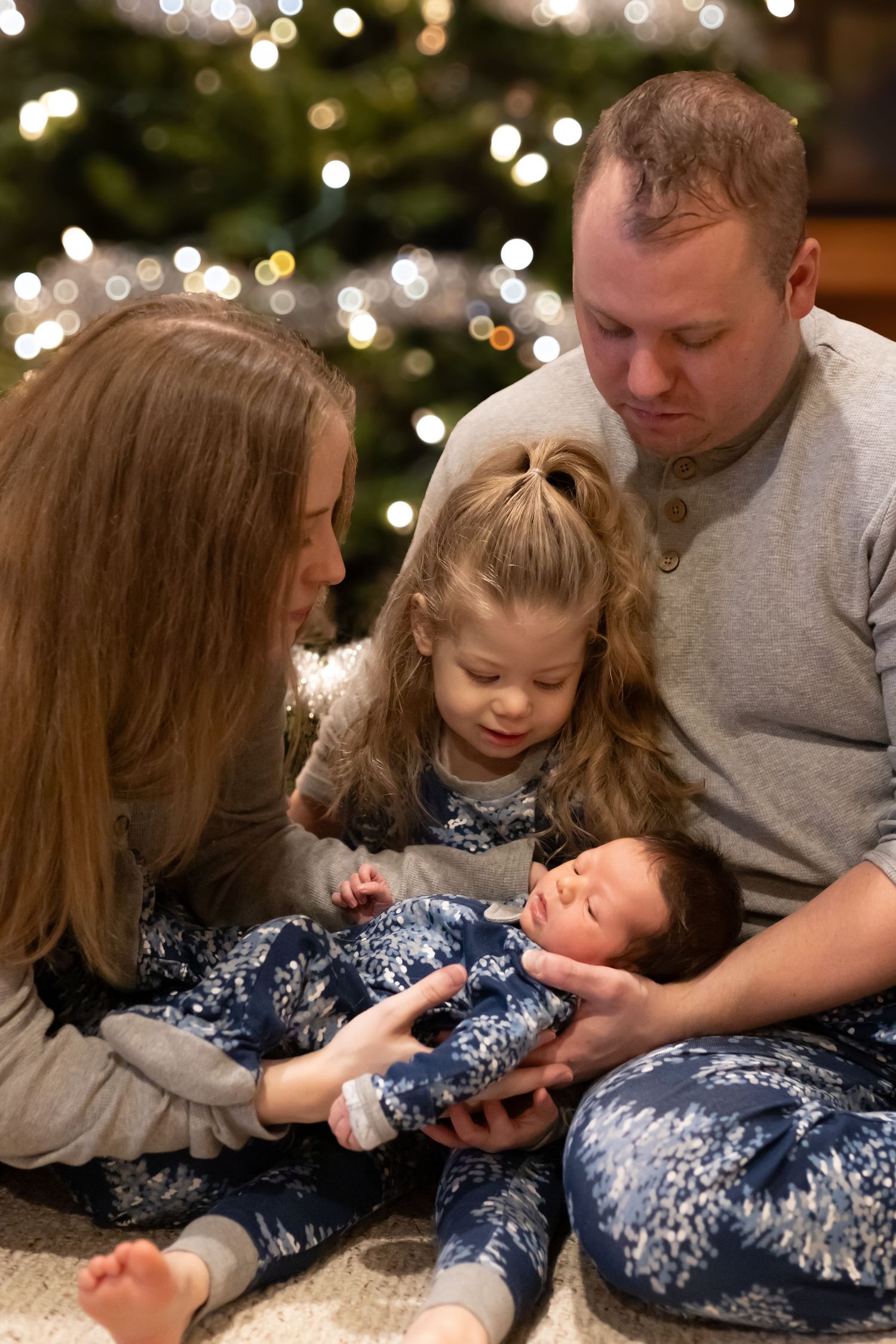 Family sitting together on floor near Christmas tree with twinkling lights in background.