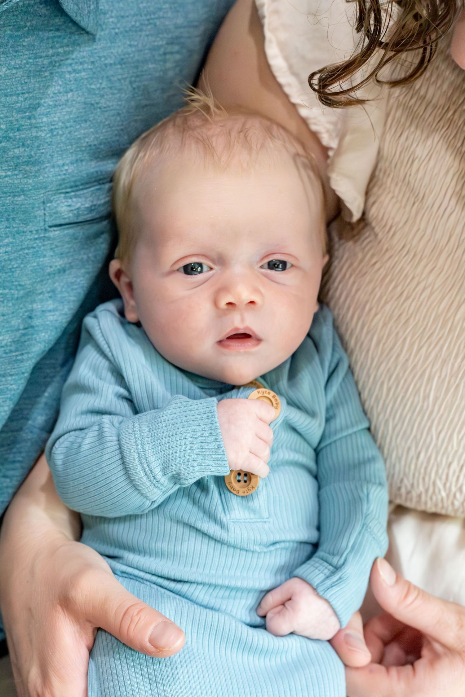 Baby wearing light blue sweater lies on cream colored fabric looking up at camera.
