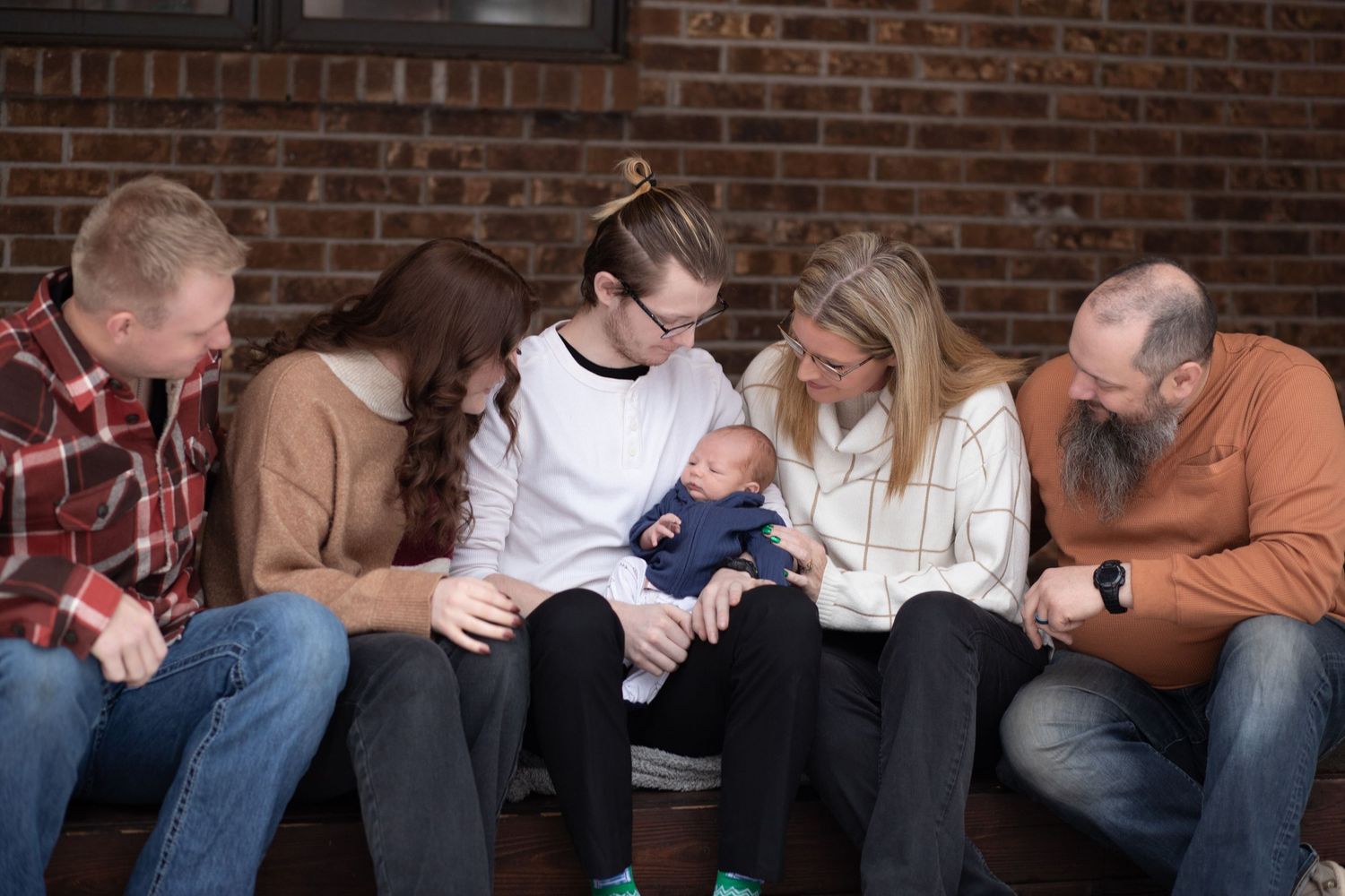 A series of people sitting on couches looking down at a baby in a brick-walled room.