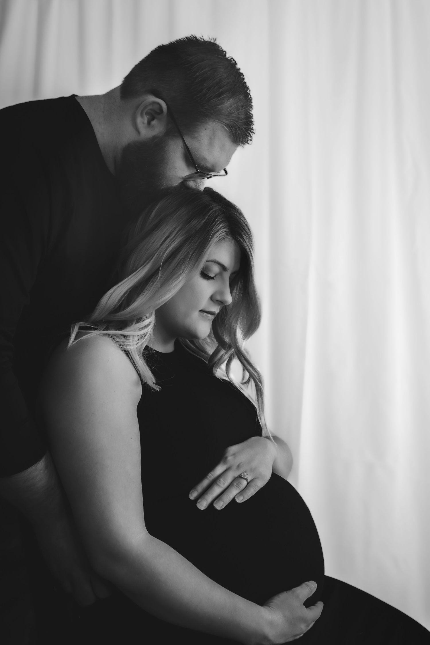 maternity photography black and white, mom is sitting in chair, black dress, dad is standing behind kissing her head