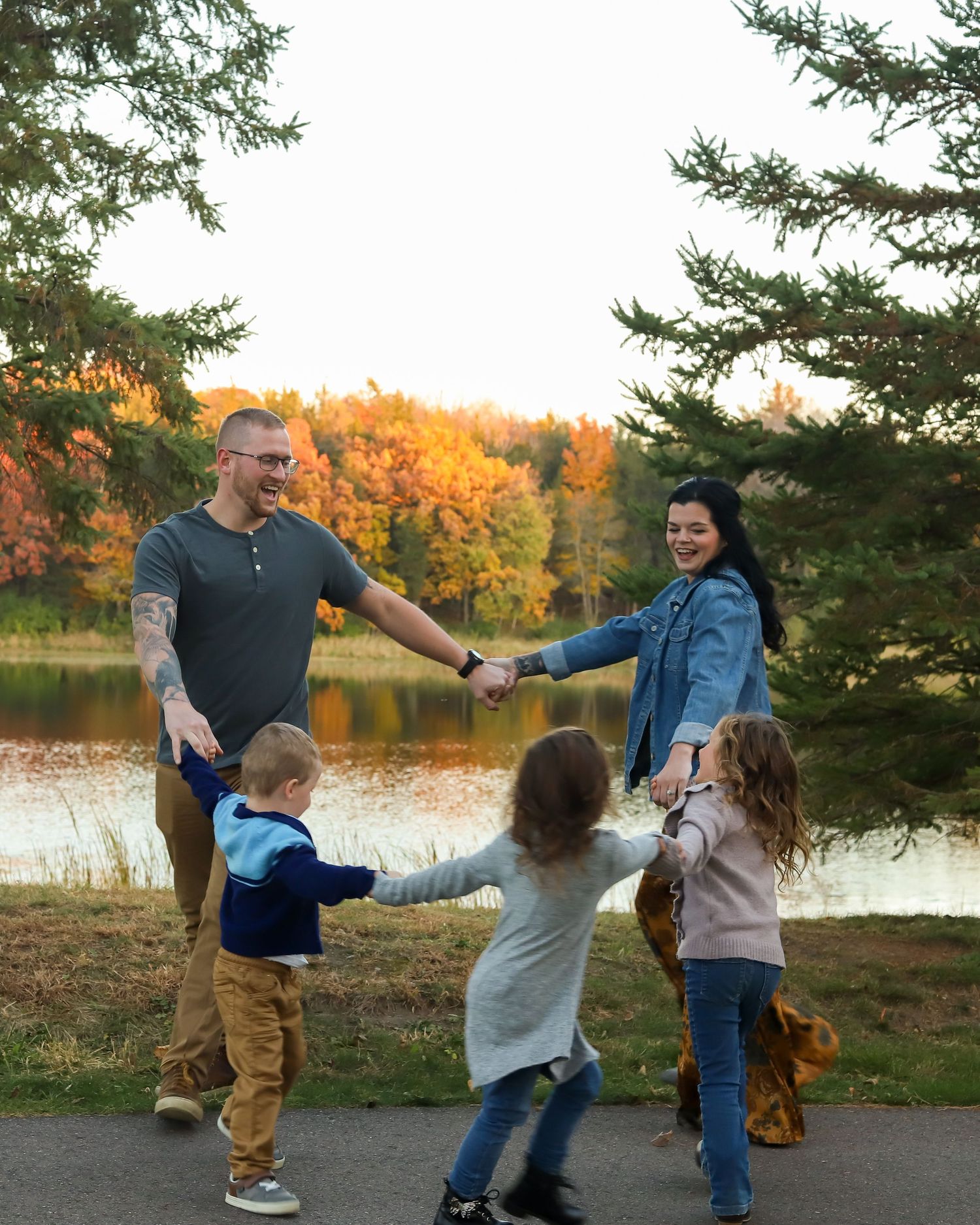 A family holds hands in a circle and dances together against a backdrop of autumn trees and a lake at sunset.