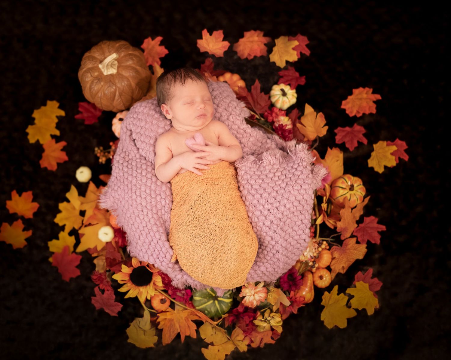 A sleeping baby in autumn-themed setting with orange pumpkin and colorful fall leaves arranged in circle on dark background.