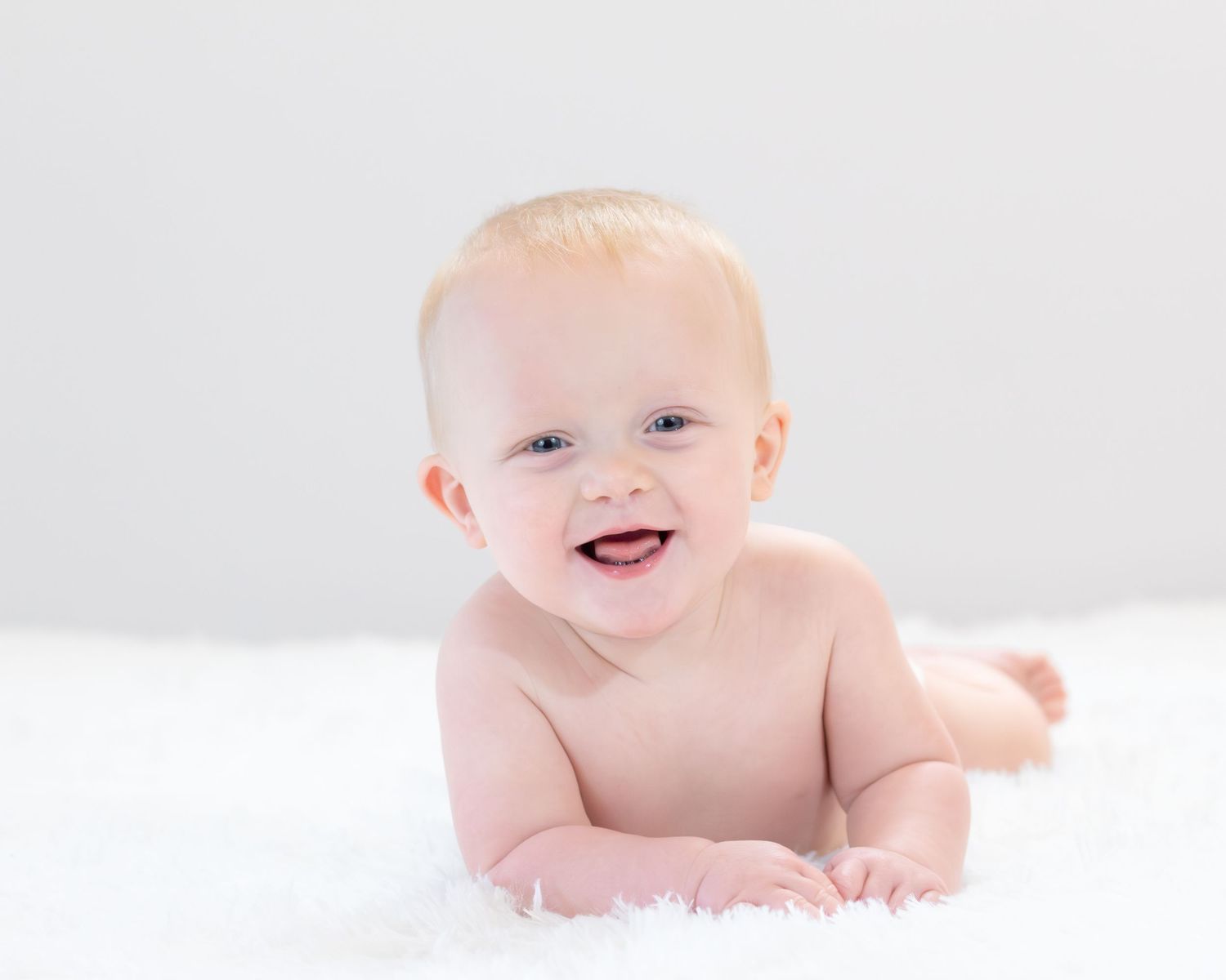 A happy infant lying on white bedding smiles brightly while looking at the camera.