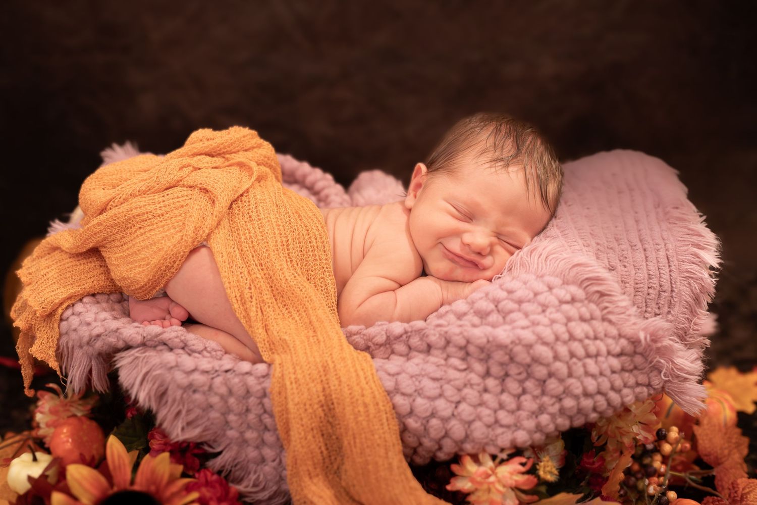 Sleeping baby wearing orange knit wrap lies on soft pink textured blanket with autumn leaves in background.