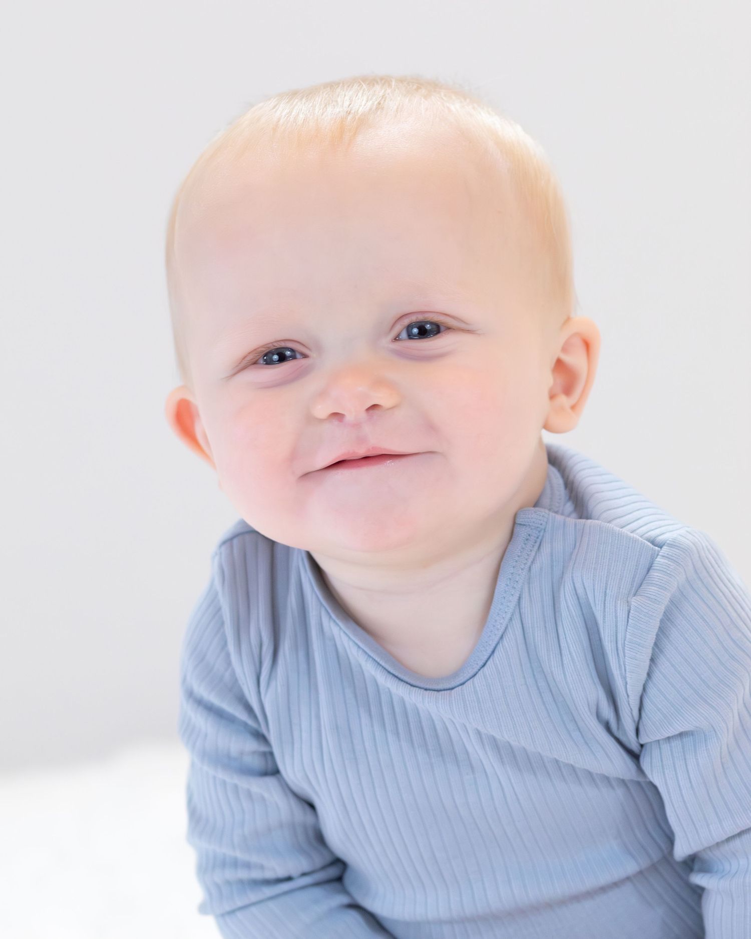 A baby in a light blue ribbed shirt smiles sweetly while lying down against a white background.