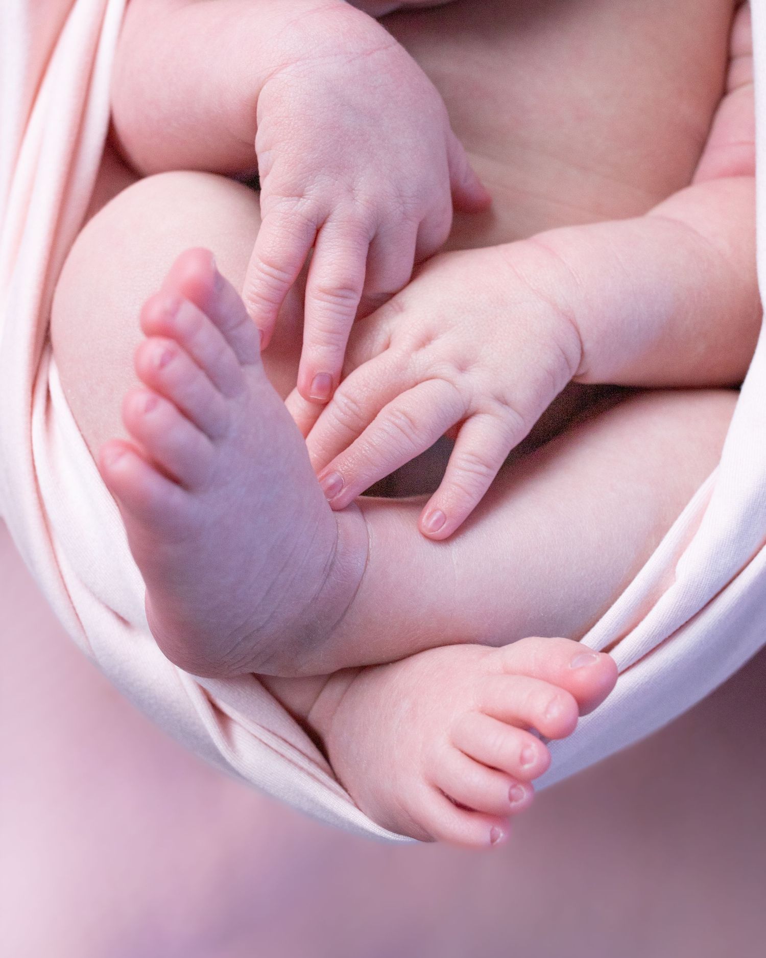 Baby's tiny feet and hands reaching against a pink background in soft lighting.