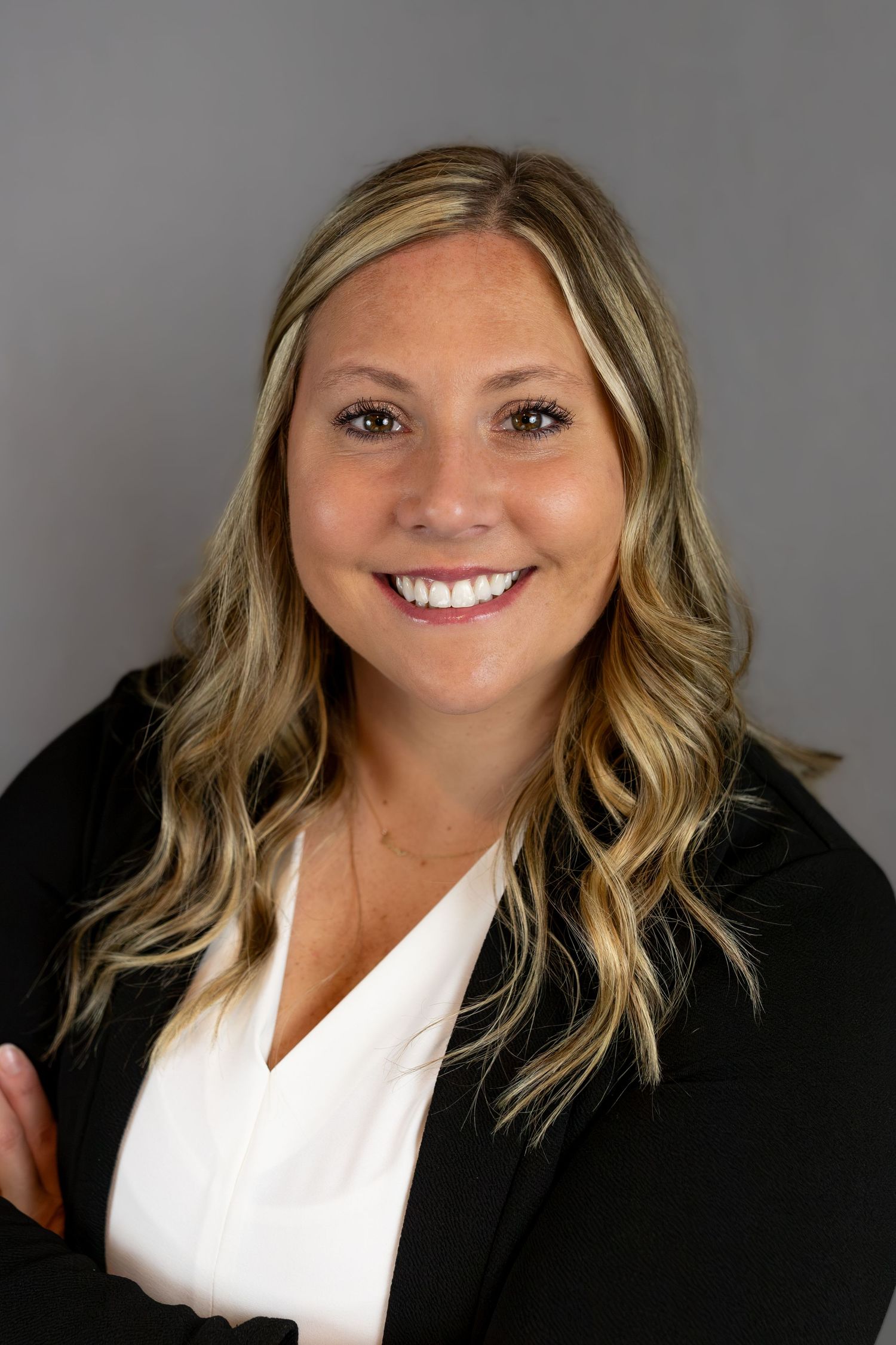 Professional headshot of a smiling blonde woman in a black blazer and white blouse against a gray background.
