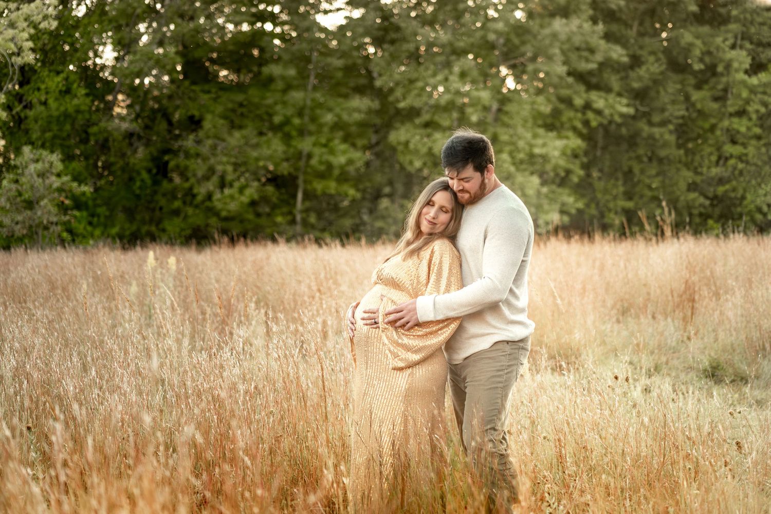 Couple embracing in golden field, pregnant woman in tan dress, man in white sweater, surrounded by tall grass and trees.