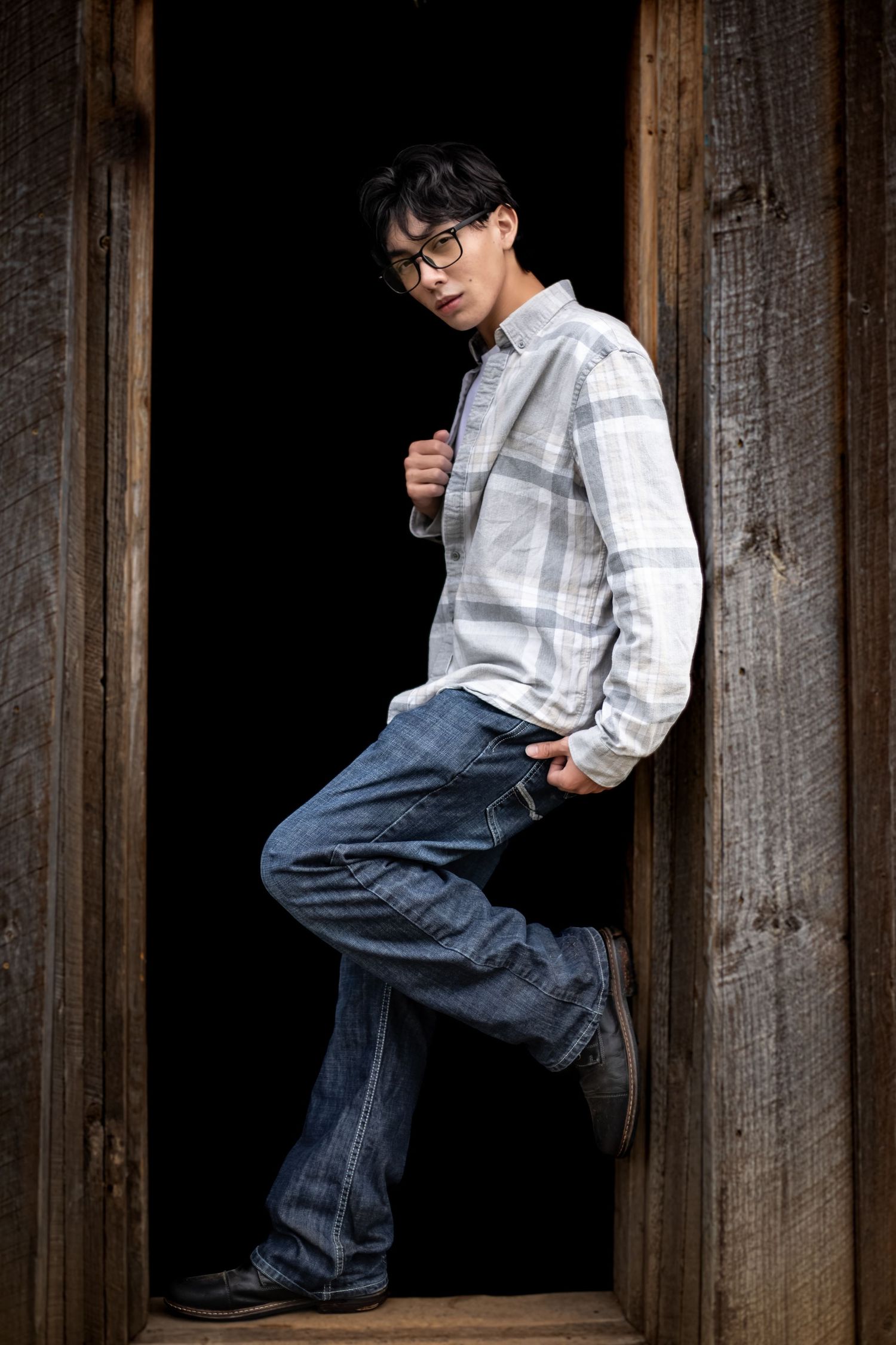 Young Asian man in plaid shirt and jeans leaning against rustic wooden doorframe with dark background.