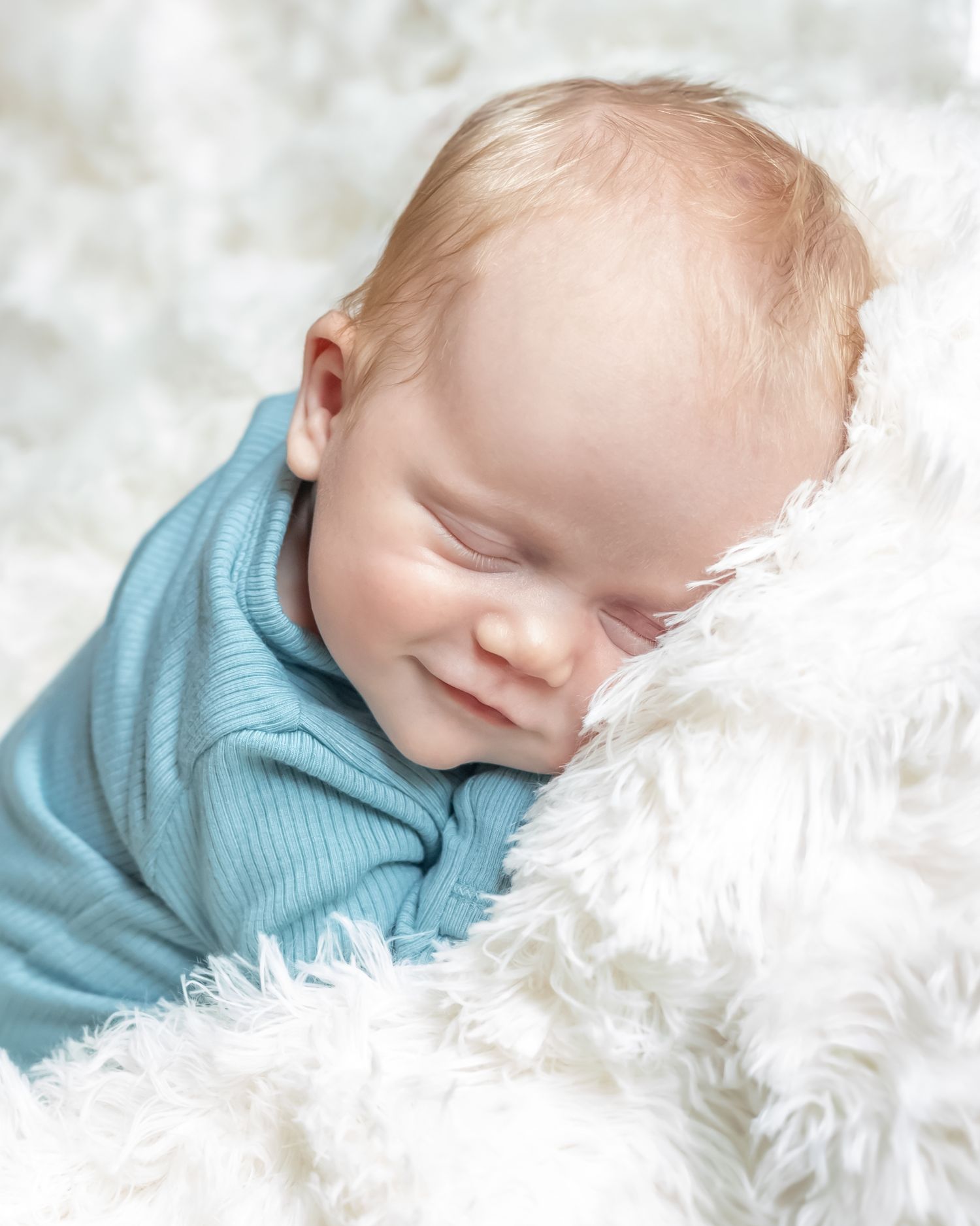 Peaceful sleeping baby wearing turquoise shirt nestled in soft white fluffy blanket.