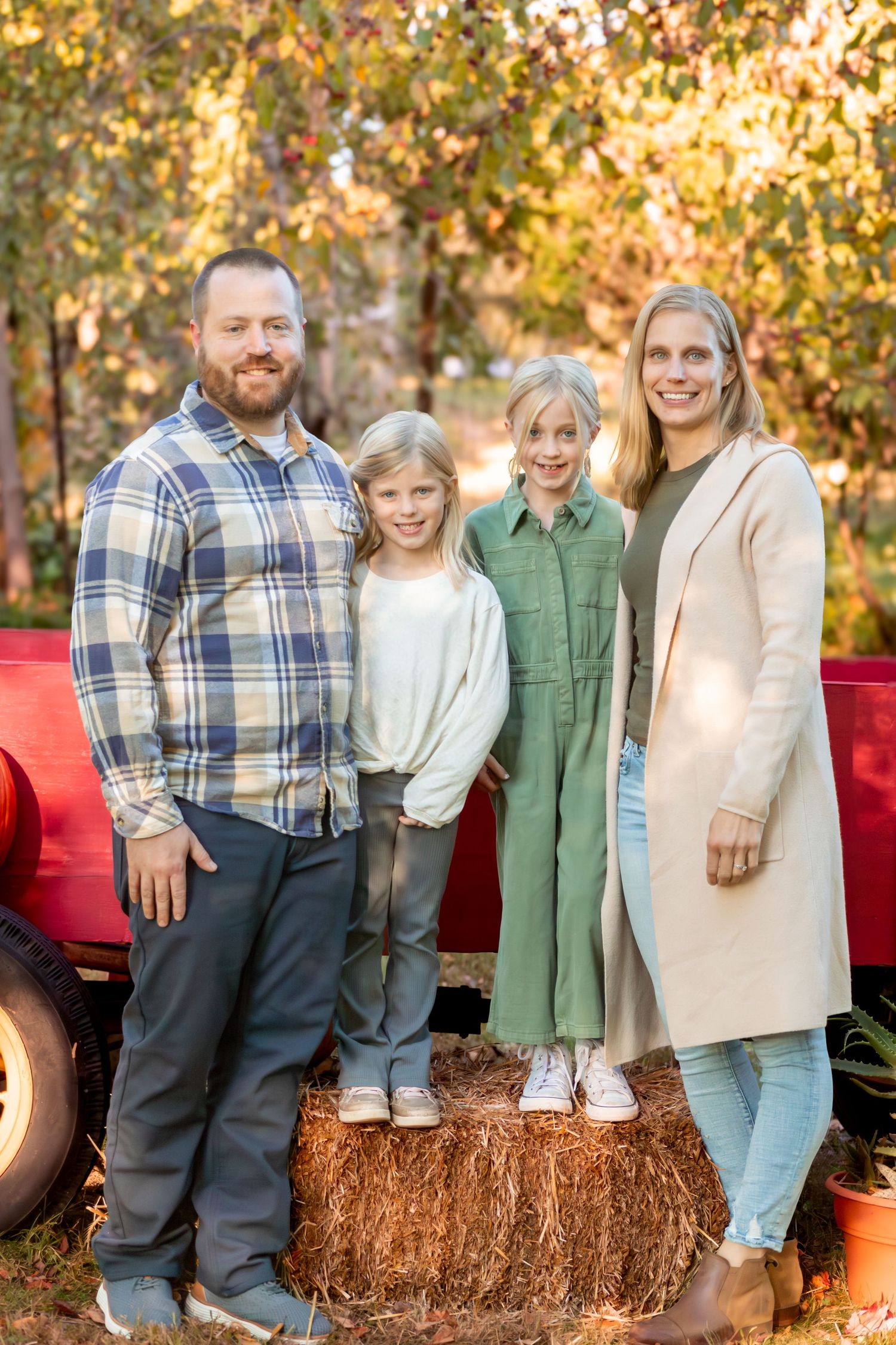 Family sitting together on hay bales during autumn with colorful trees behind them.