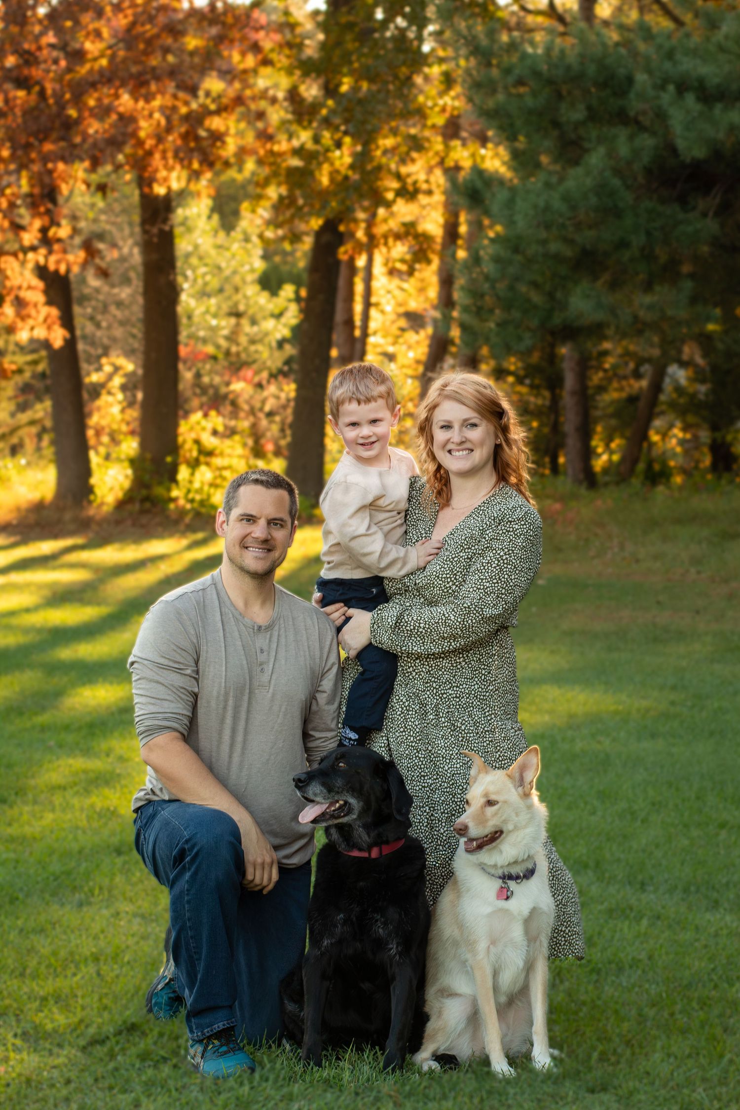 Family portrait with two dogs on grass with autumn trees in background.
