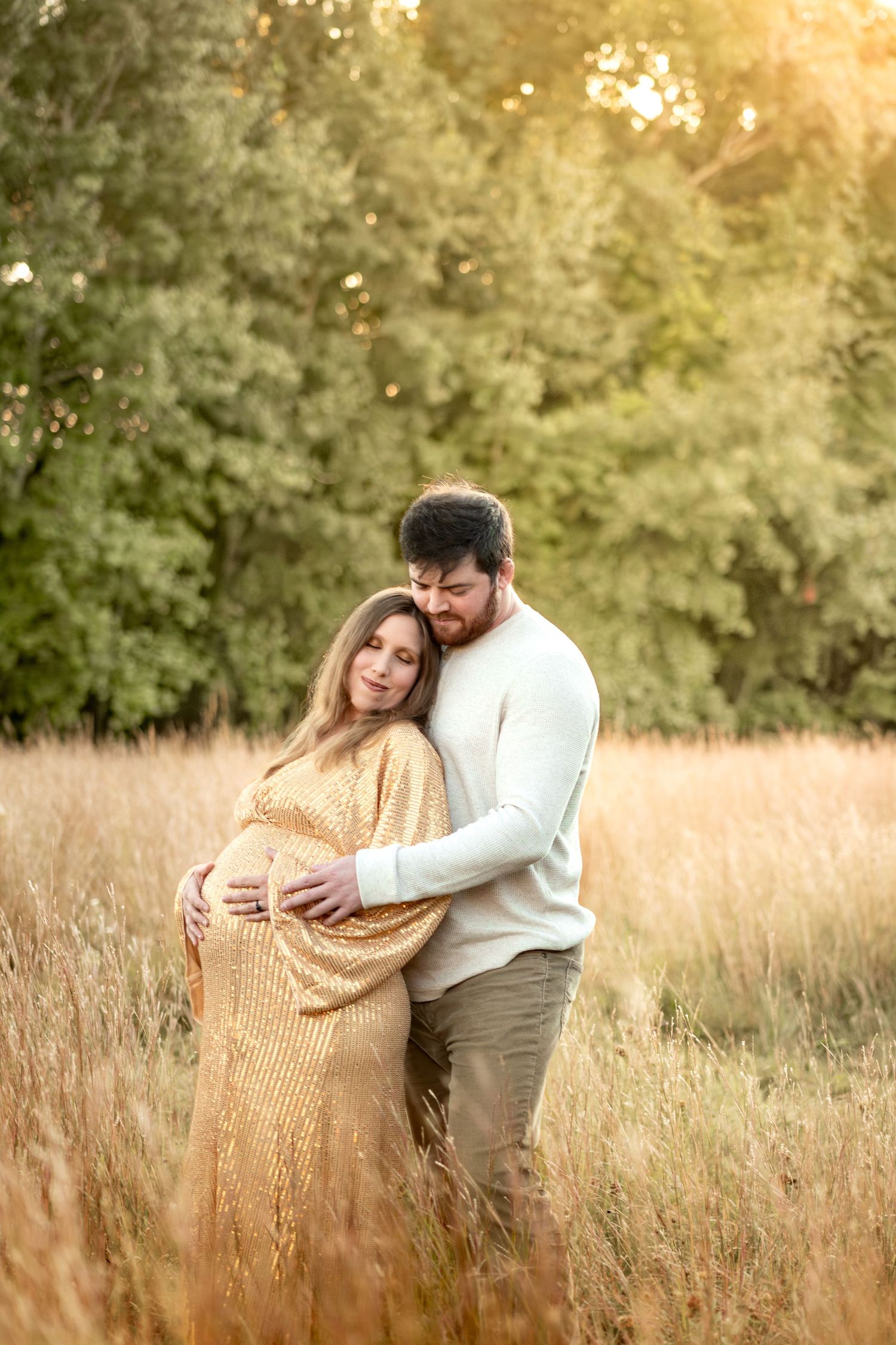 Intimate maternity photo of couple embracing in a sunlit meadow with golden grass.