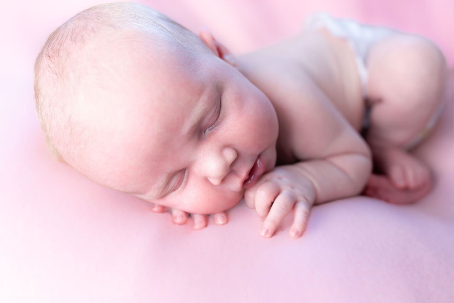 A peaceful sleeping baby curled up on soft pink blankets in a gentle newborn pose.