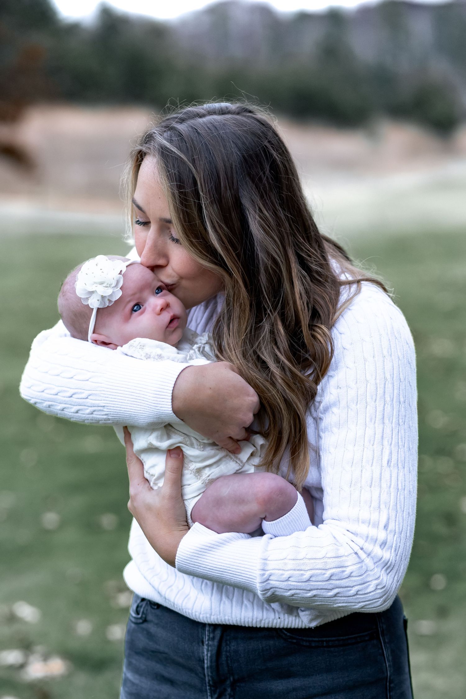A mother in a white sweater tenderly embraces and kisses a newborn baby in a series of intimate outdoor portraits.