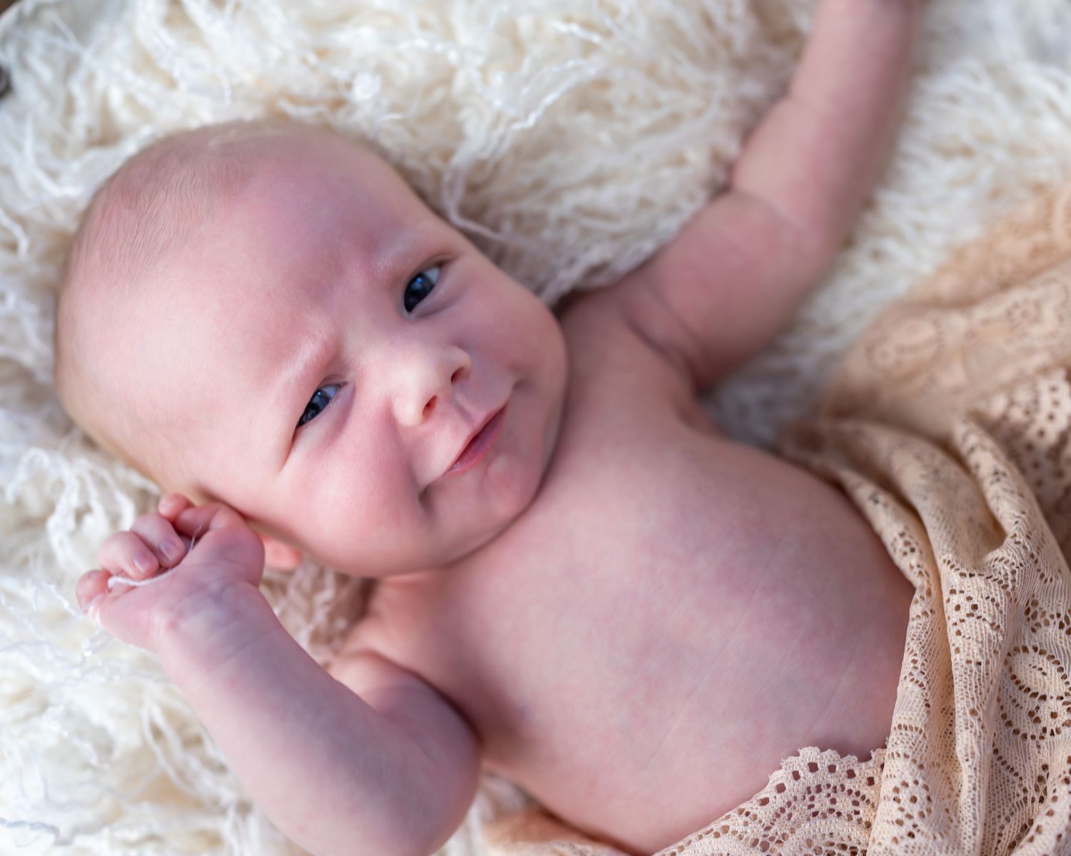 A sweet newborn baby lying on soft white lace fabric looking up at the camera with bright eyes.
